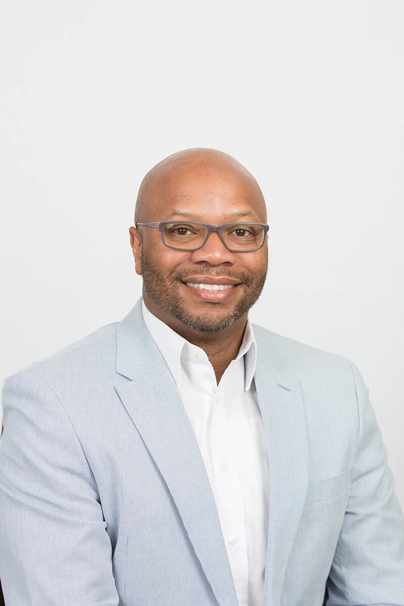 Headshot of a smiling man with glasses, wearing a light-colored blazer and white shirt against a plain white background.