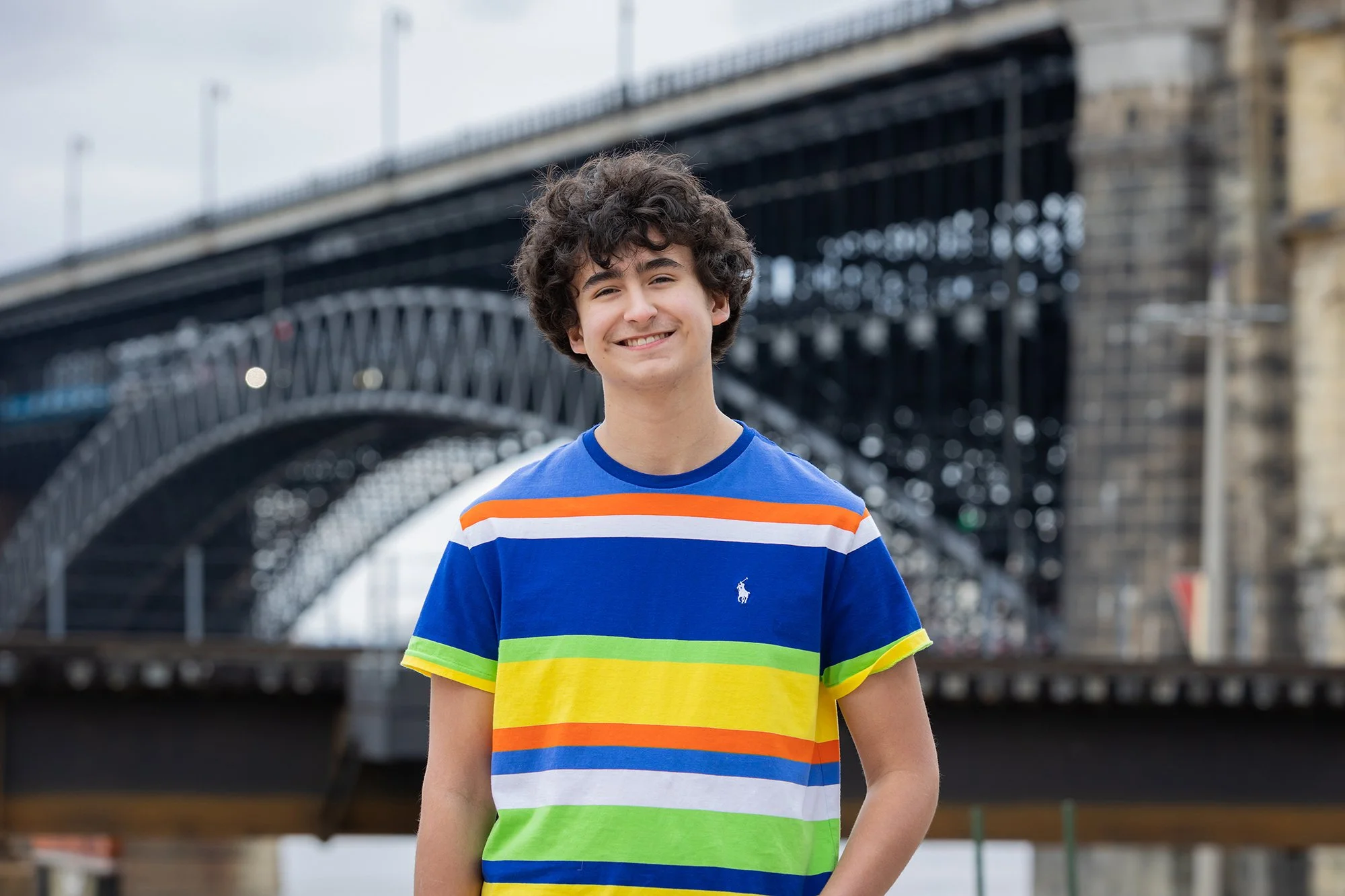 A young boy with curly hair smiling, wearing a rainbow-colored striped t-shirt, standing in front of a bridge.