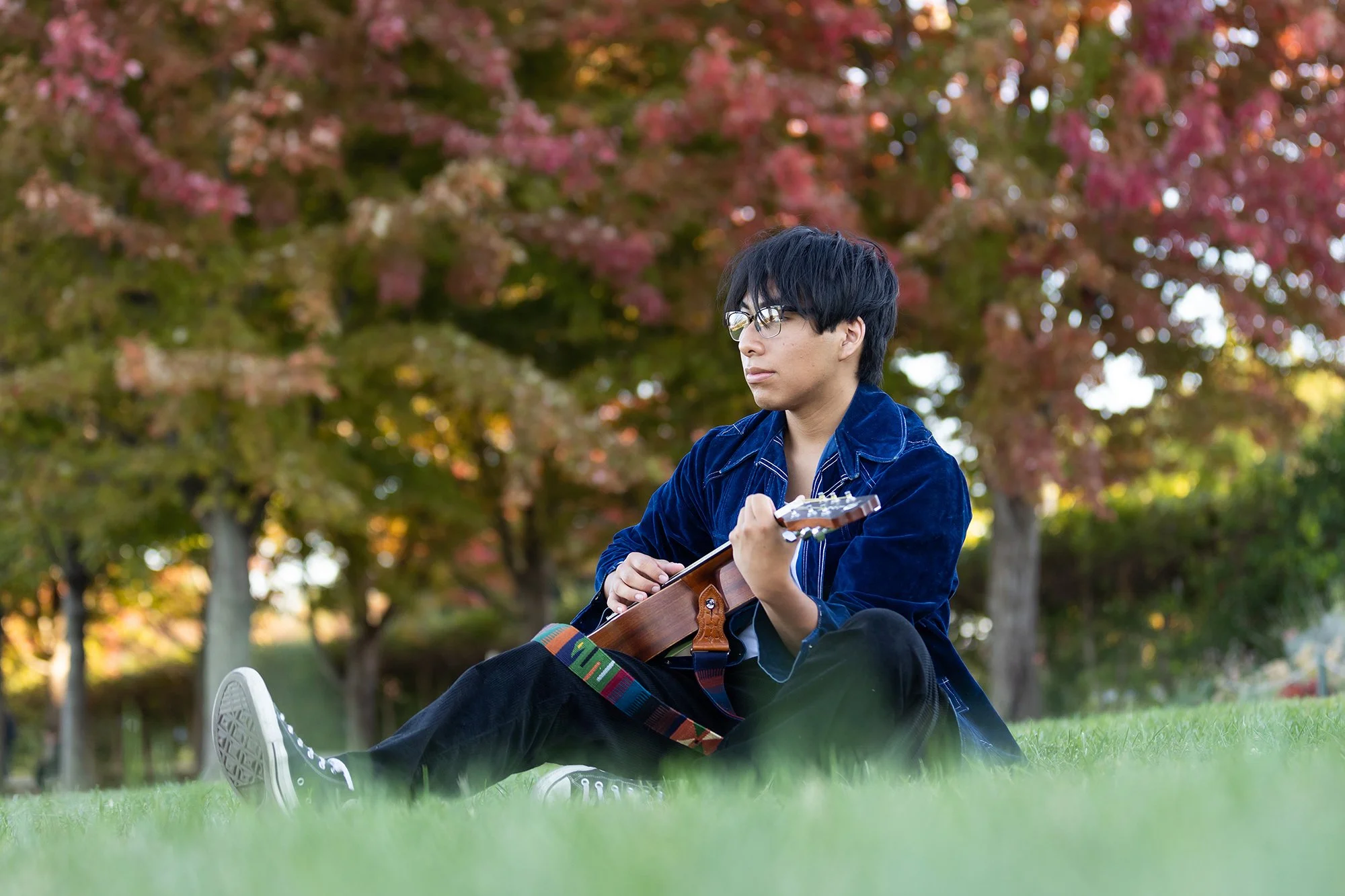 Young man with black hair, glasses, wearing a blue jacket, sitting on grass and playing guitar outdoors with autumn trees in the background, posing for his senior portraits.