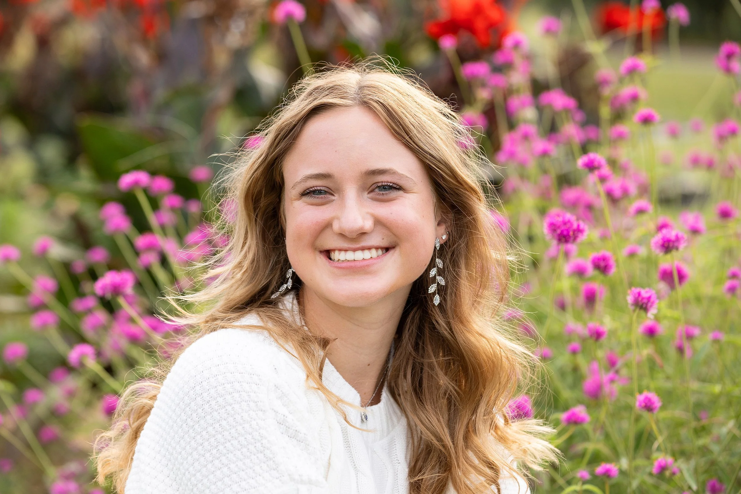A young woman with long, wavy blonde hair, smiling in a garden with pink flowers and greenery in the background.
