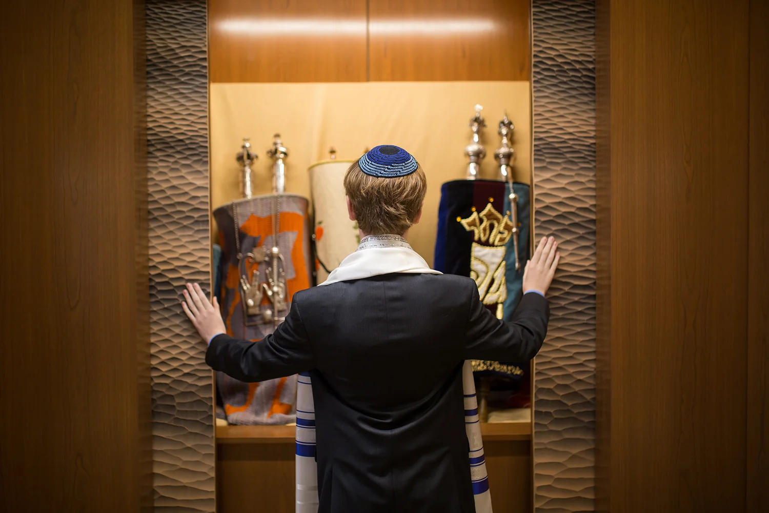 Person dressed in traditional Jewish prayer attire, wearing a kippah, praying before the Torah in a synagogue.