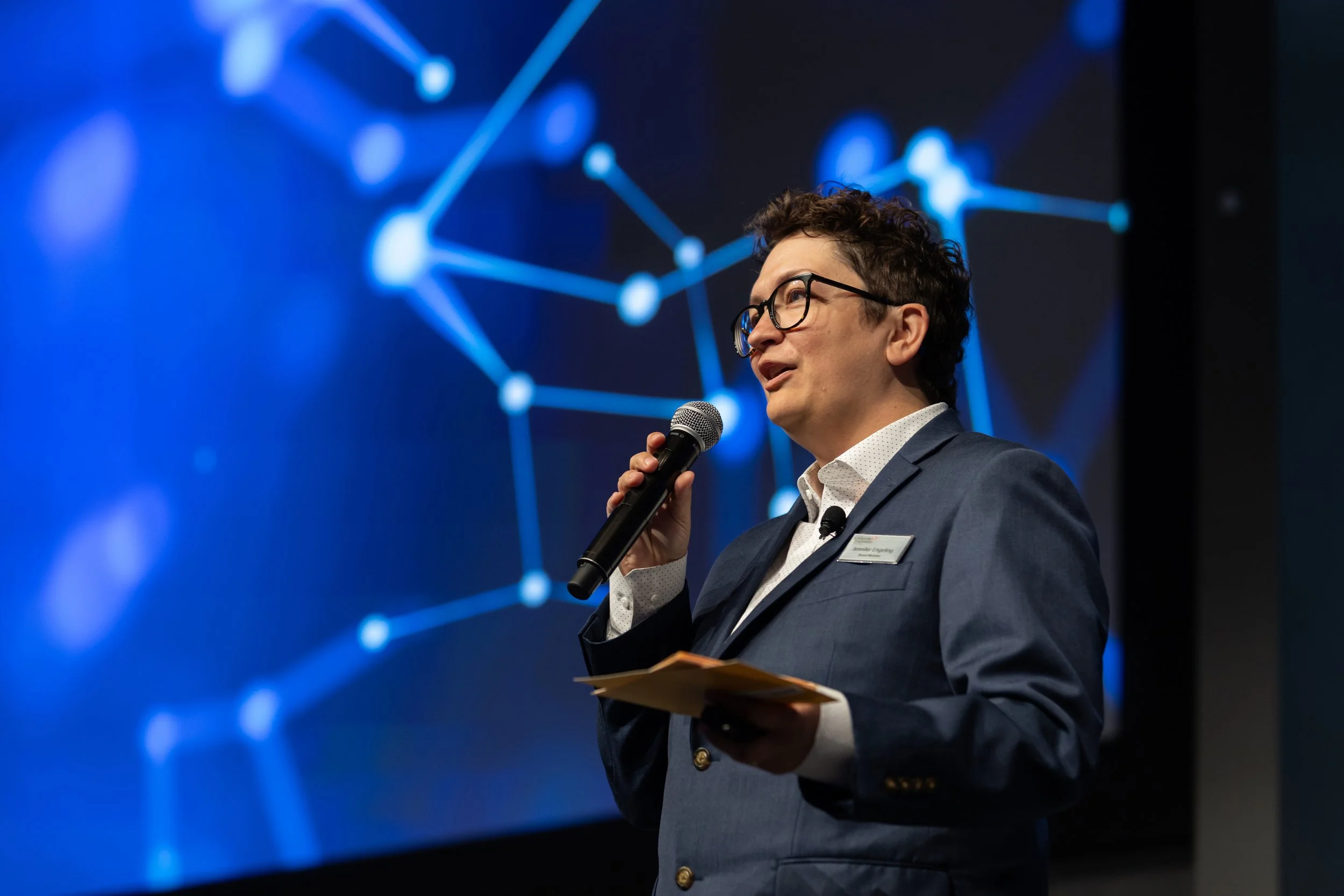 A man wearing glasses and a suit is speaking into a microphone on stage. He is holding a notepad and standing in front of a digital projection of a blue interconnected network graphic.