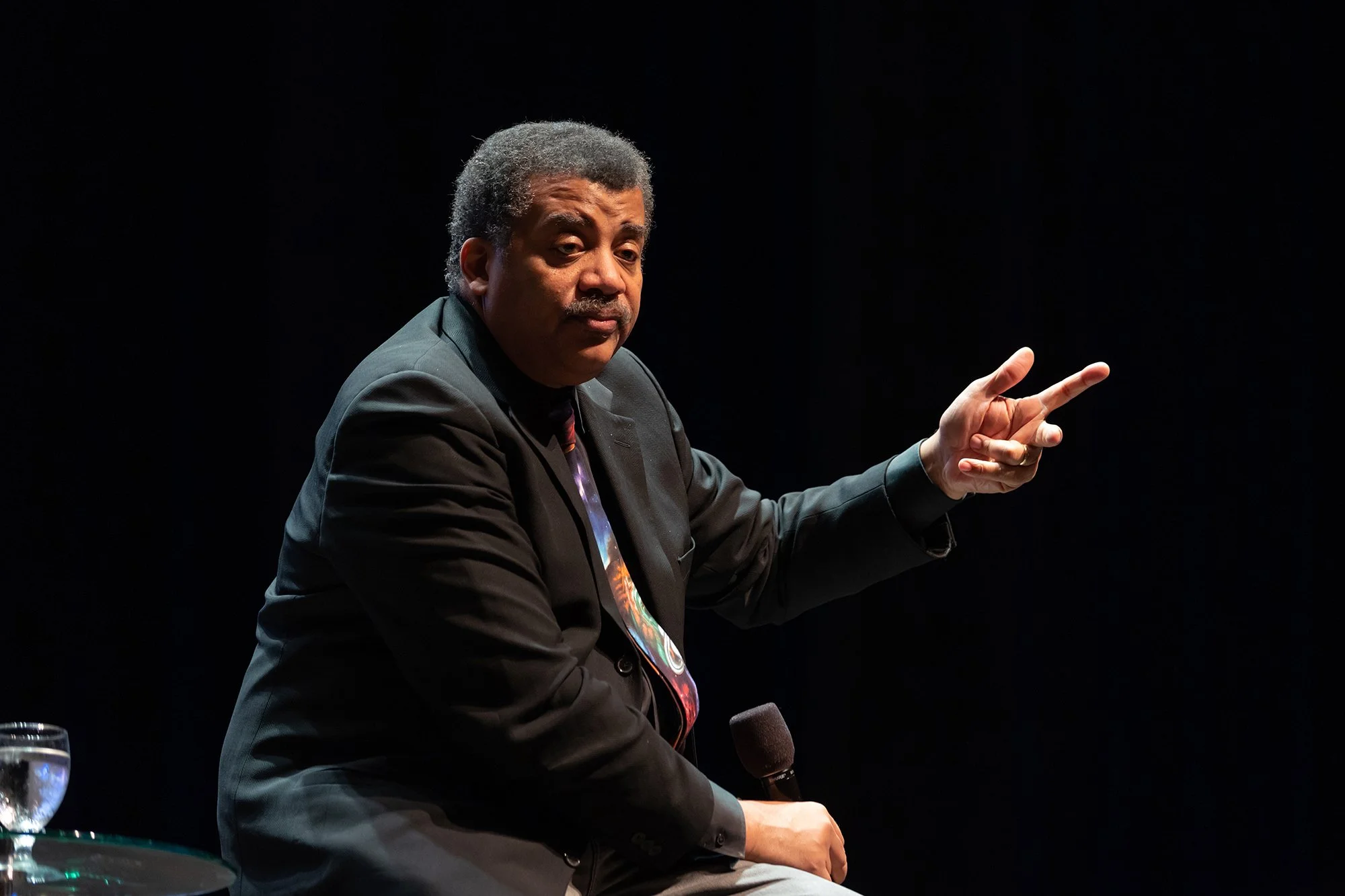 A man with curly hair and a mustache sitting on a stage, wearing a dark suit, holding a microphone, and pointing with his right hand against a dark background.