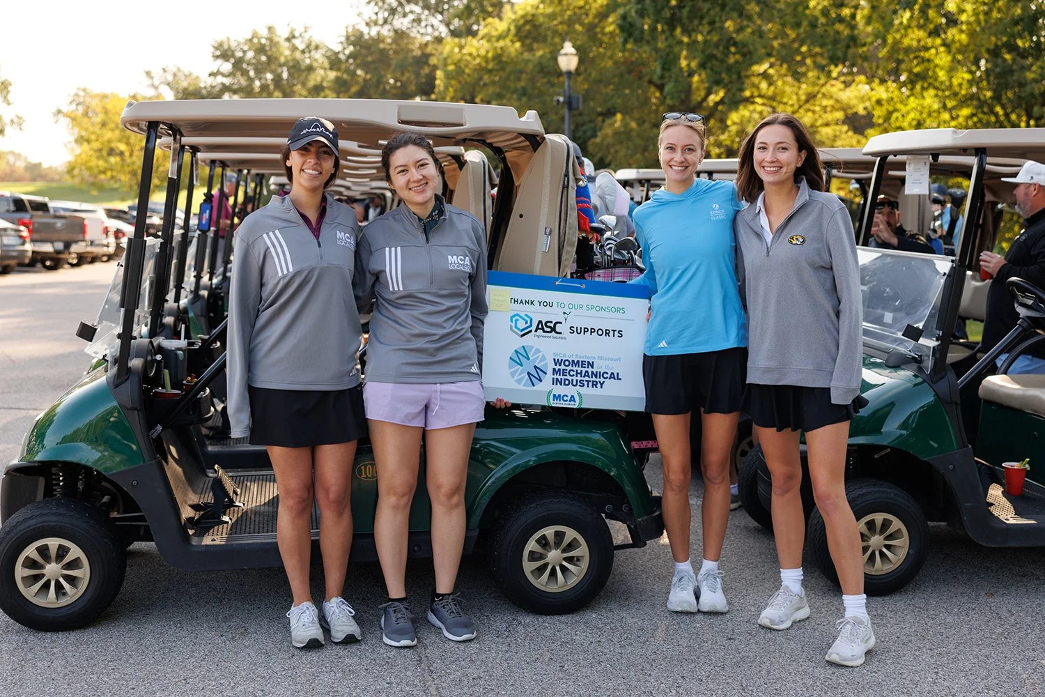 Four women standing in front of golf carts at a golf course, smiling. One cart displays a sign thanking sponsors for supporting the Missouri Women in the Mechanical Industry. Three women are wearing casual athletic clothing, and one is wearing a blue