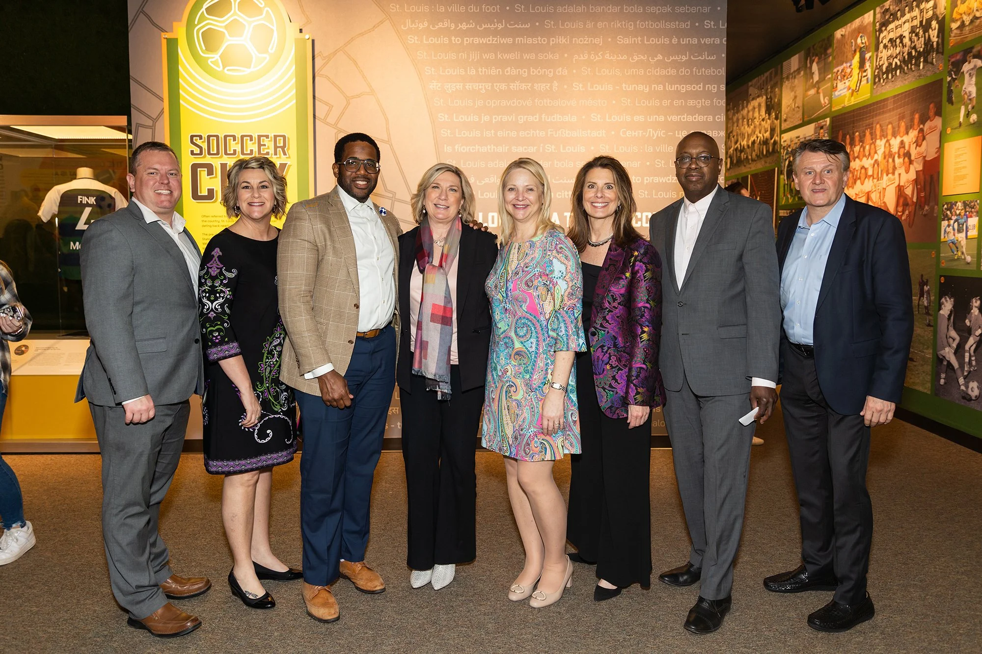 A diverse group of nine people standing together indoors at a sports exhibit, smiling at the camera, with a yellow and green soccer-themed display and photos of soccer players on the wall behind them.