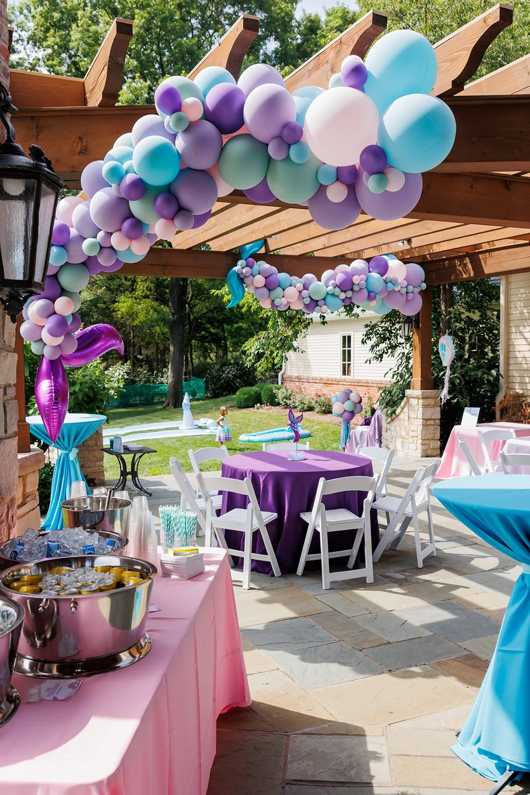 Decorated outdoor patio with pastel-colored balloons in purple, blue, pink, and white forming an arch and garlands. Tables with purple and pink tablecloths, white chairs, and a table with drinks and snacks. A purple dolphin-shaped balloon and a small