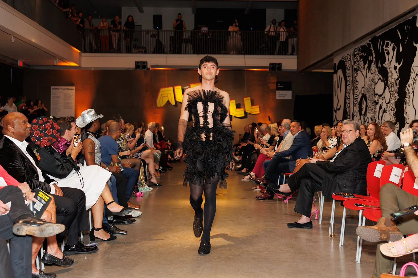 A fashion model walks down the runway at a fashion show, wearing a black dress with feather and lace details. The audience is seated on both sides, watching the show with some taking photos.