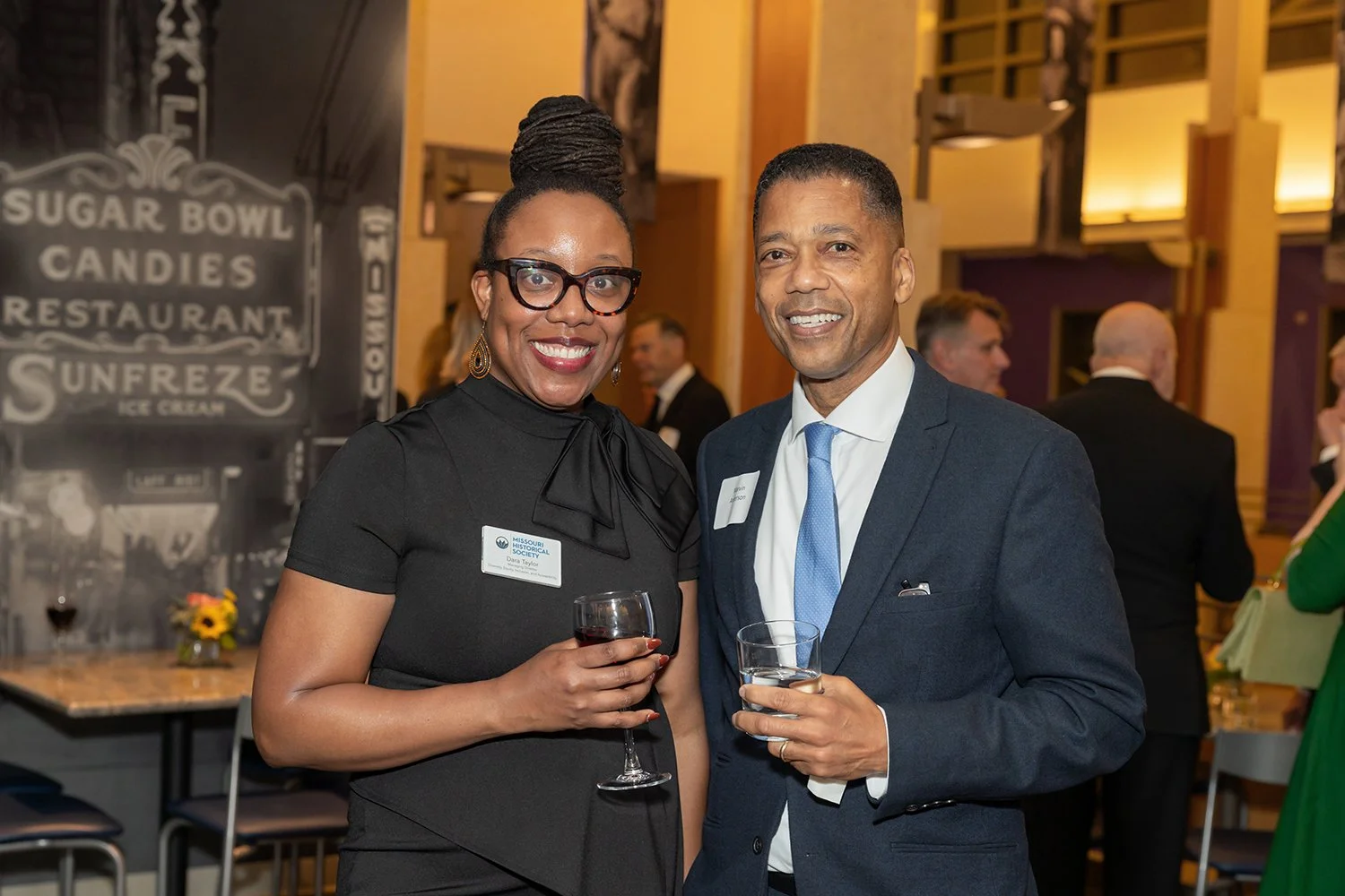 A woman and a man at a formal event, smiling and holding drinks, with other guests in the background.