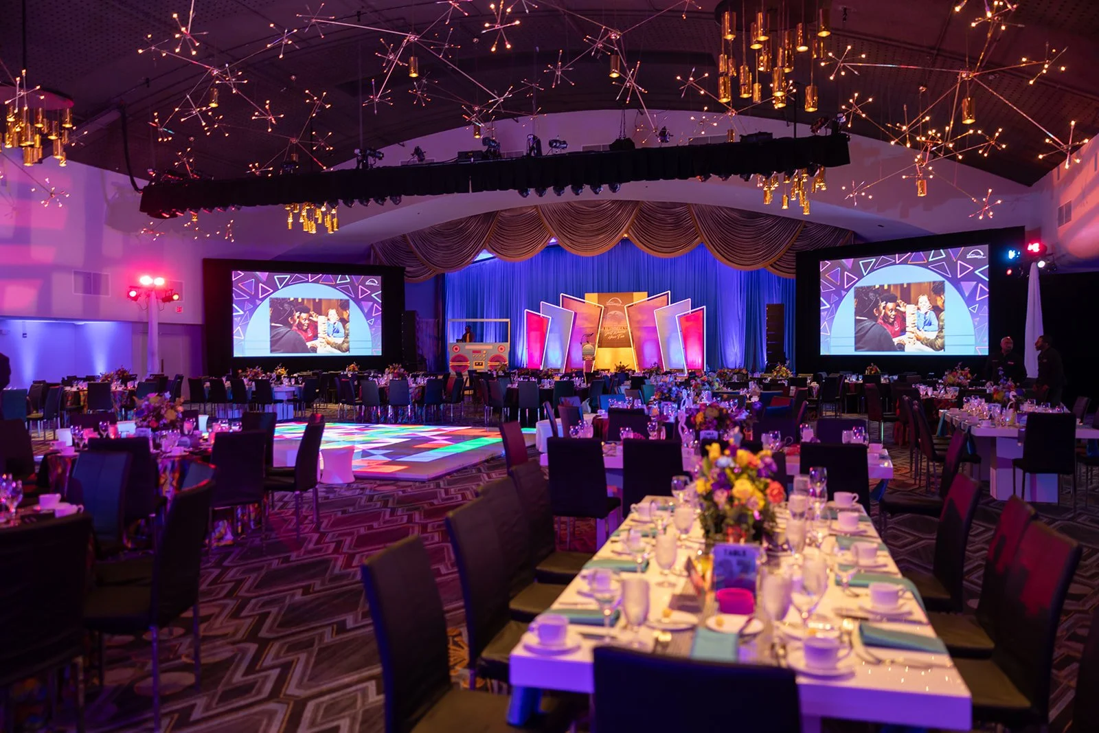 An event hall decorated for a celebration with a stage, large screens, colorful lighting, and tables set with flowers and dinnerware.