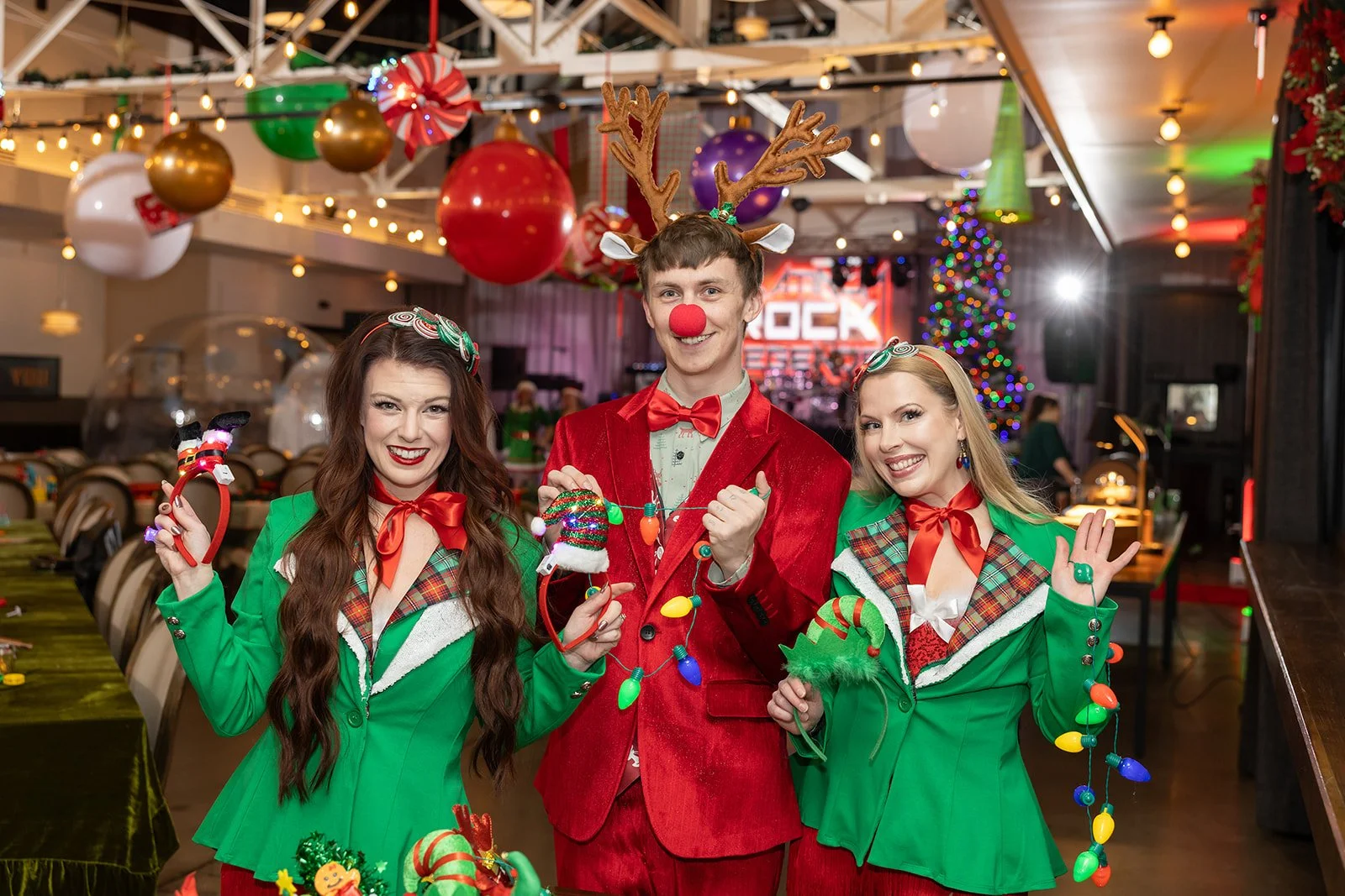 Three people dressed in Christmas-themed costumes posing and smiling in a decorated venue with Christmas ornaments, lights, and a Christmas tree.