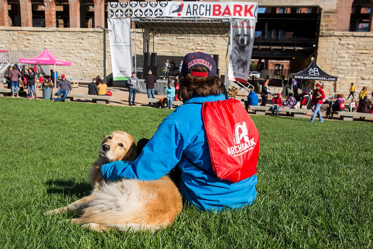 Person sitting on grass with a golden retriever at an outdoor event, with a stage and banners in the background.