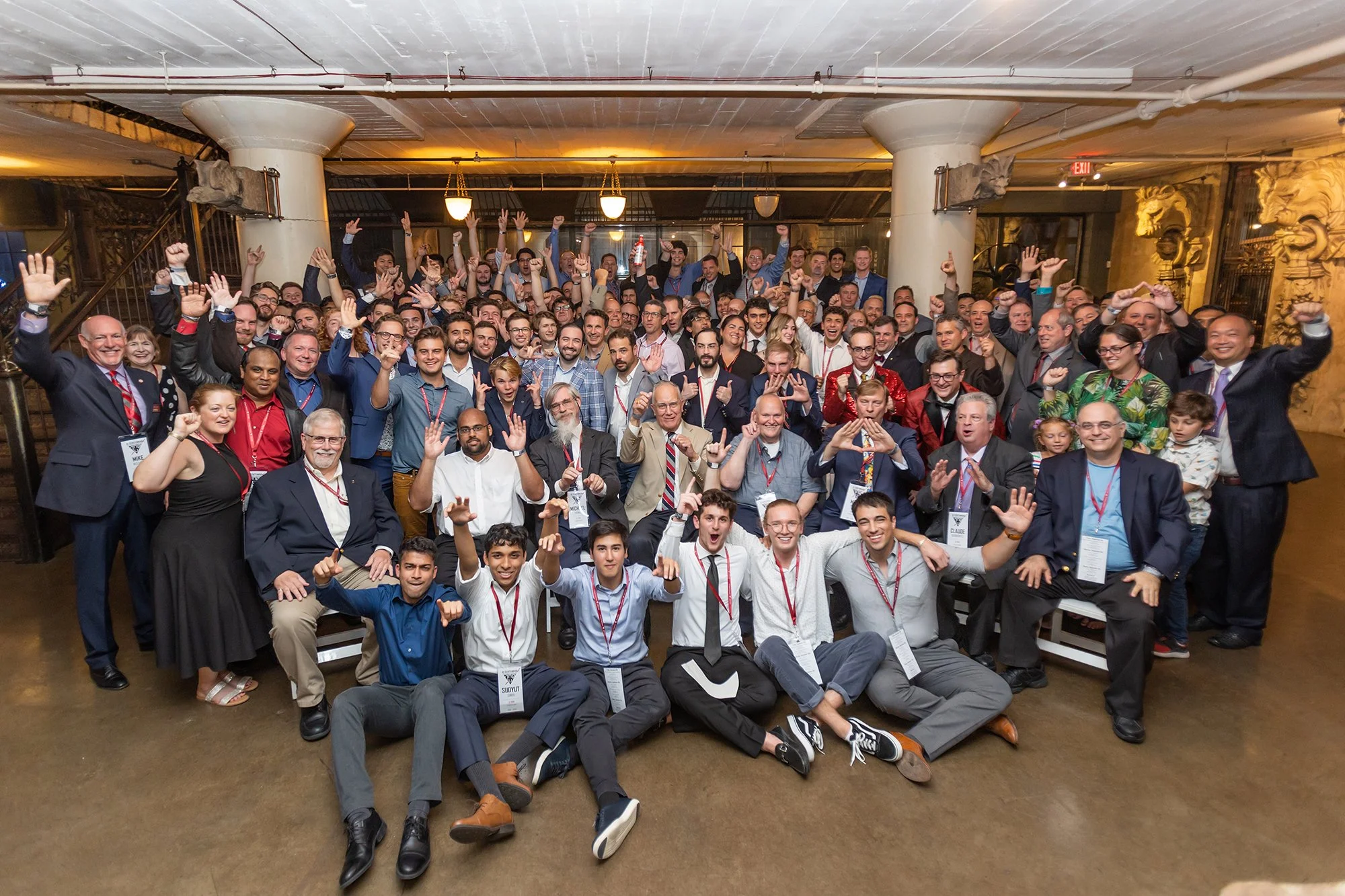 Group of diverse people at a conference or gathering, smiling and raising their hands, in an indoor venue with warm lighting and ornate decorations.