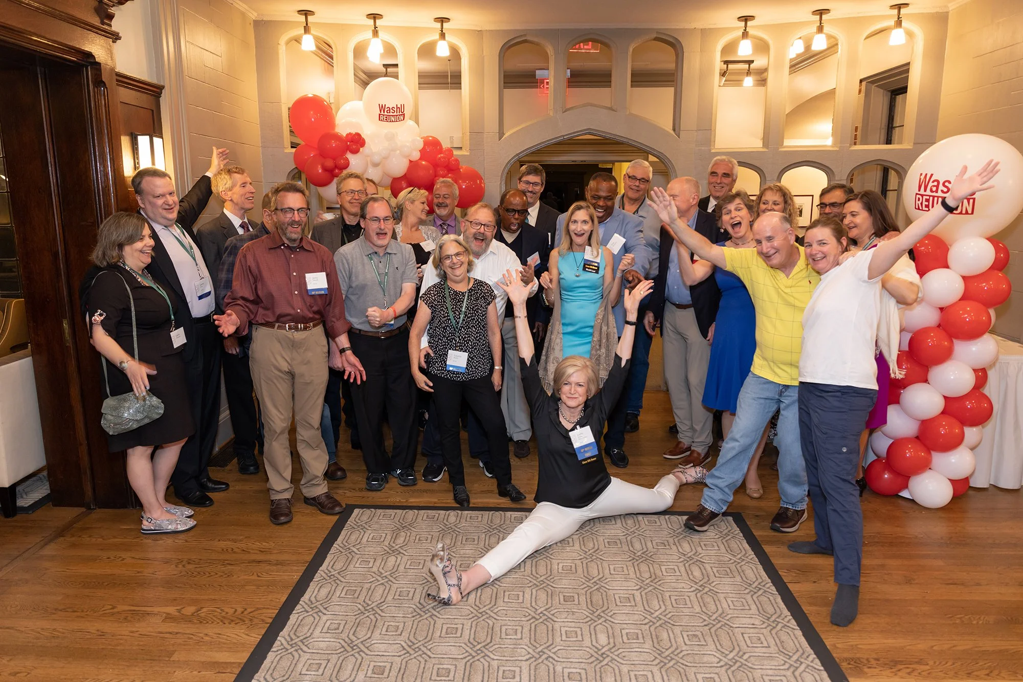 A large group of people at a reunion event, some are smiling, talking, and cheering. There are balloons with the words "Washl REUNION" on them, and the setting appears to be a banquet hall or restaurant with warm lighting and decorative arches.