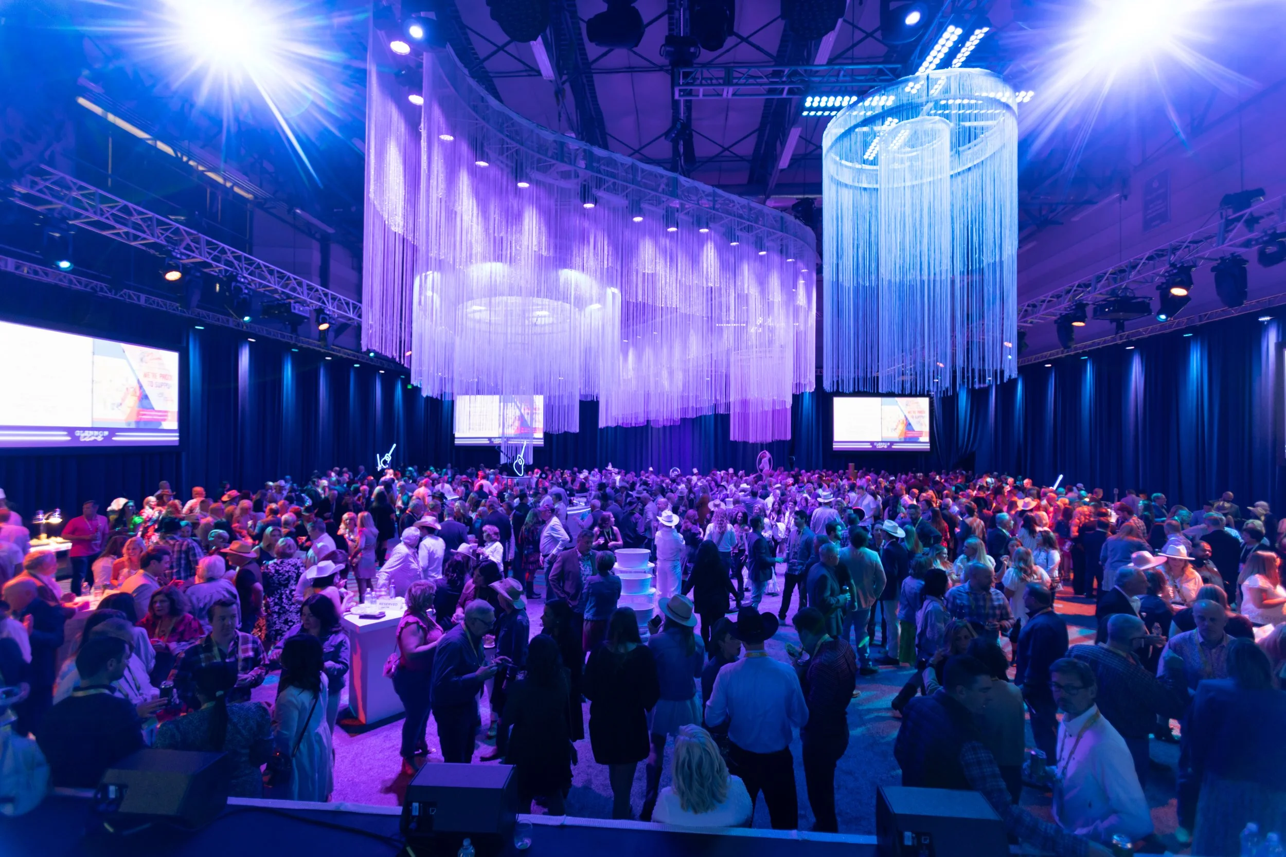 Large indoor event with many people, illuminated by purple and blue lights, featuring hanging chandeliers and multiple screens on the walls.