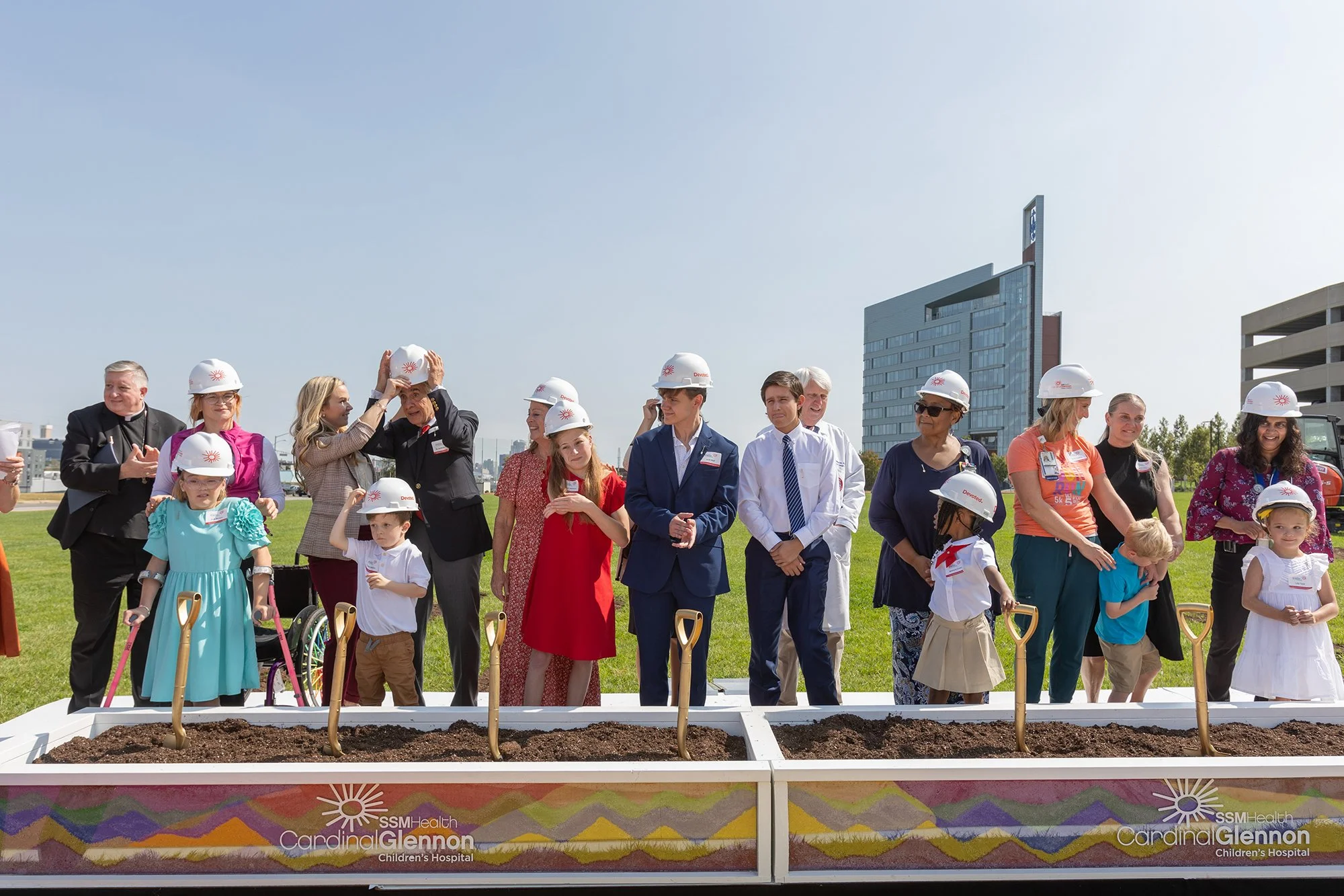 A group of people, including children and adults, wearing construction helmets, standing outdoors in front of two planters with soil and shovels for a groundbreaking ceremony, with a cityscape in the background.