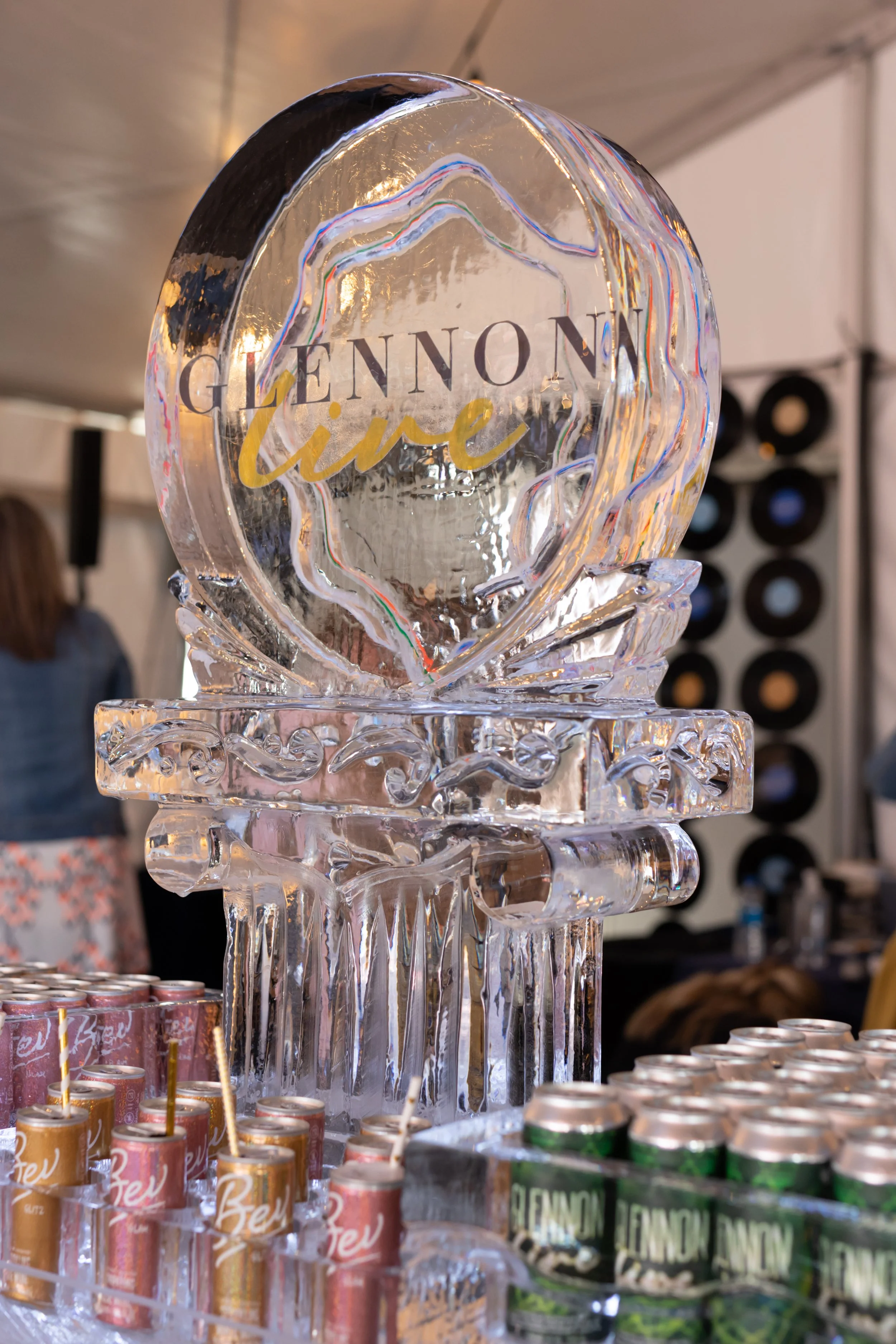 Ice sculpture with the text 'Glennon Live' at an event, with cans of beverages in front.