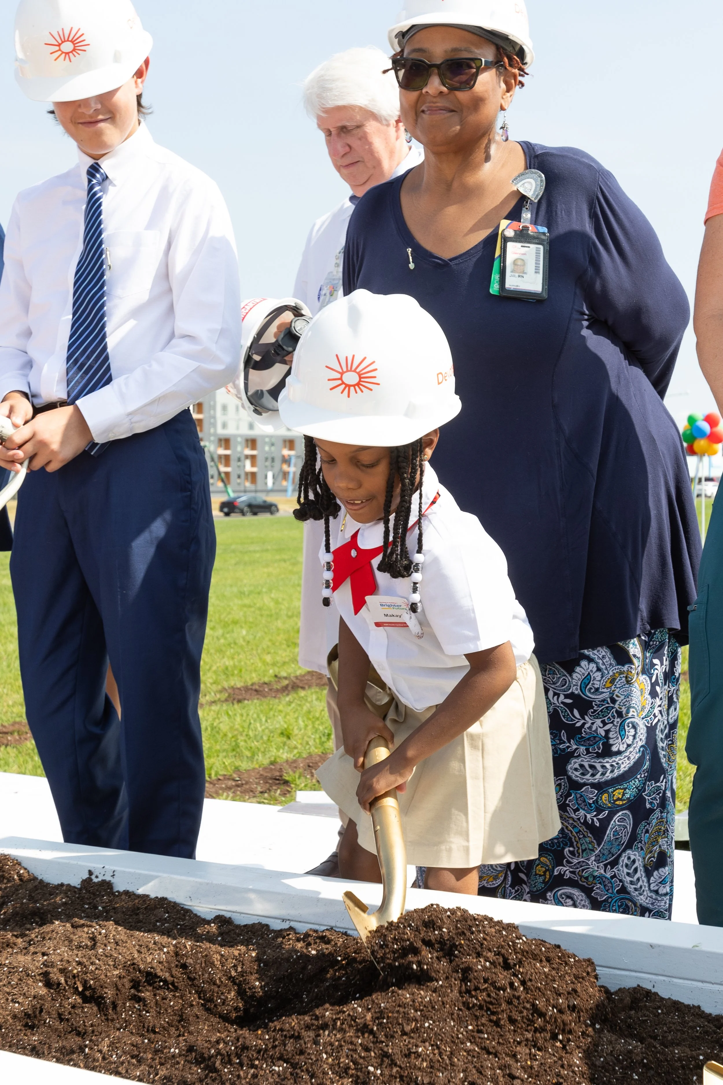 A young girl in a white uniform and a white hard hat is digging in a garden bed with a small shovel while people around her, including a woman in sunglasses and a man in a white shirt, watch during a community planting event.