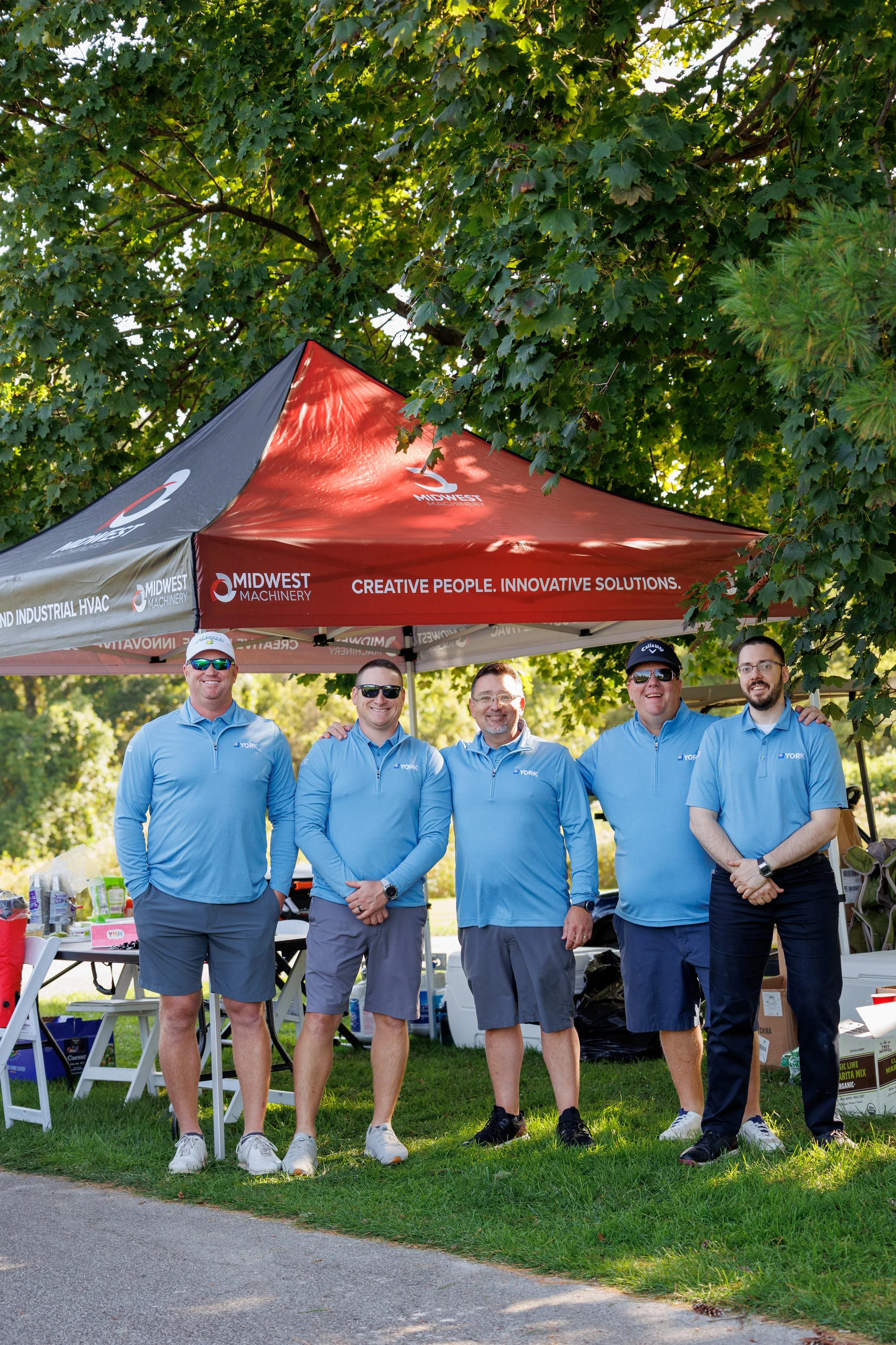 Five men standing under a Midwest Machinery tent at an outdoor event, smiling and posing for a photo on a sunny day.