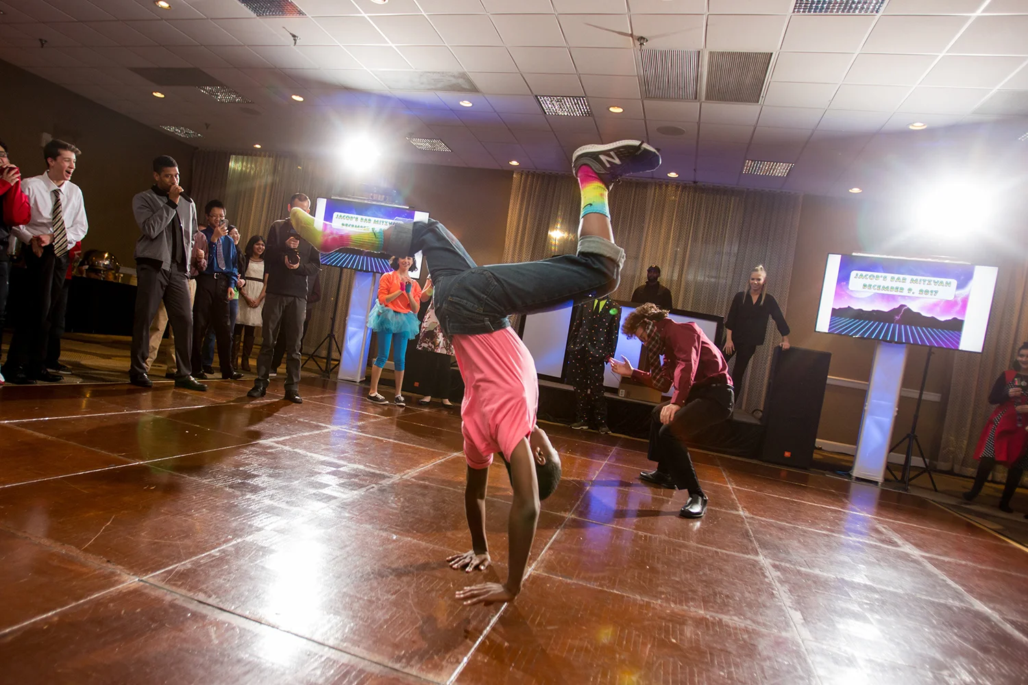 Person performing a headstand or break dance move on a wooden dance floor at a party, with several onlookers watching, some clapping, and event screens displaying 'Jace's Bar Mitzvah December 8, 2019' in the background.