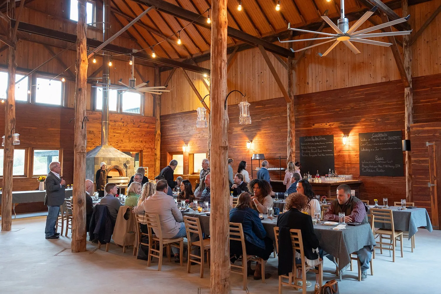 People dining at a rustic wooden banquet hall with high ceilings, large windows, string lights, and chalkboard menus on the wall.