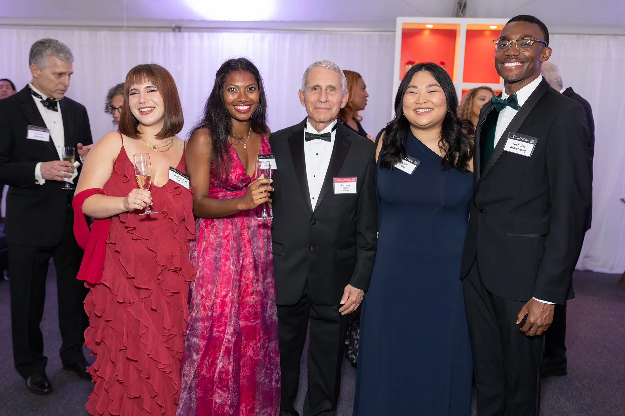 Group of five diverse people dressed in formal attire at an event, smiling and holding champagne glasses, with other guests in the background.