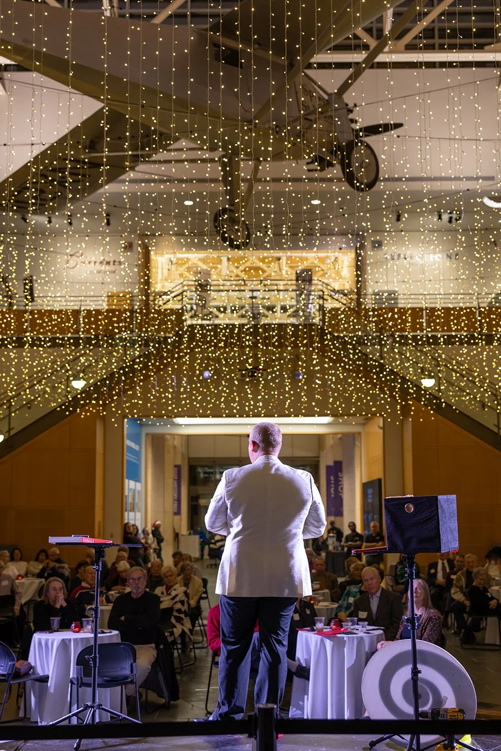 A man in a white blazer speaking to an audience at an event, with fairy lights hanging above and a small airplane model hanging from the ceiling.
