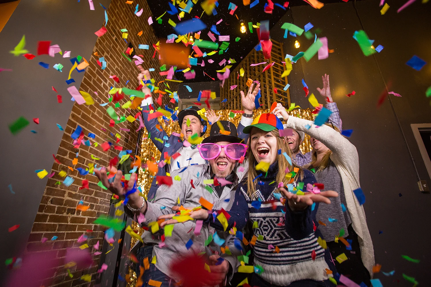 People celebrating with confetti falling, wearing party hats and funny glasses, smiling and having fun in a festive setting.