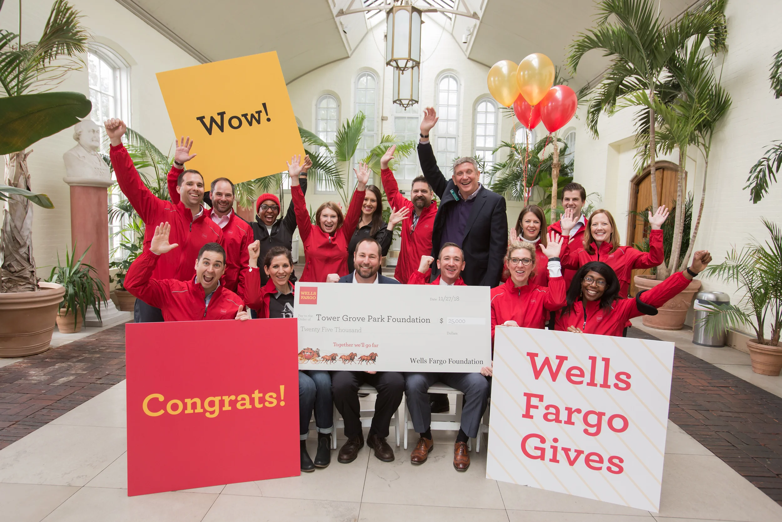 Group of people celebrating indoors with signs and a large check, wearing red jackets, some raising their hands and smiling.