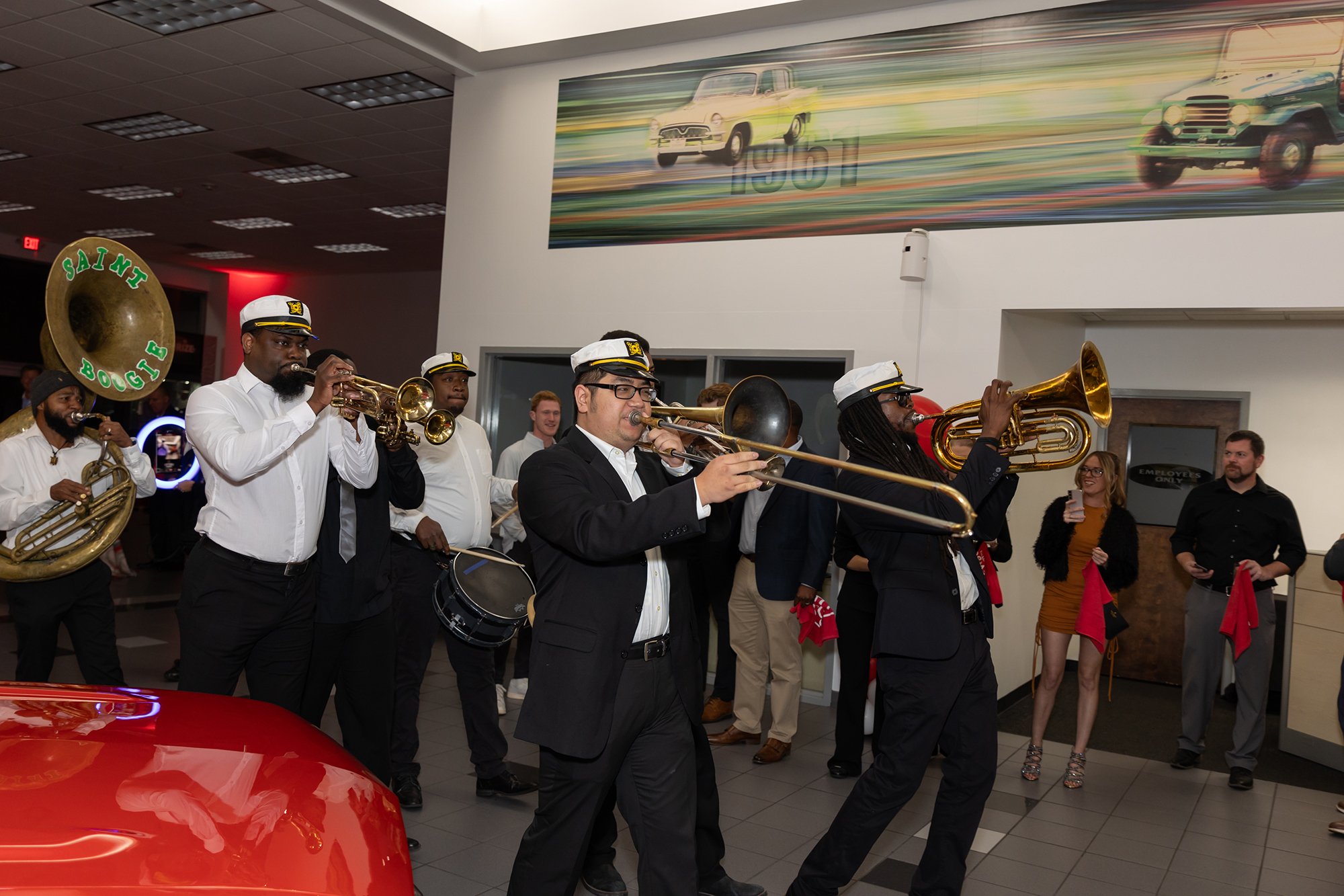 Musicians in uniform playing brass instruments at an indoor event, with onlookers in the background.