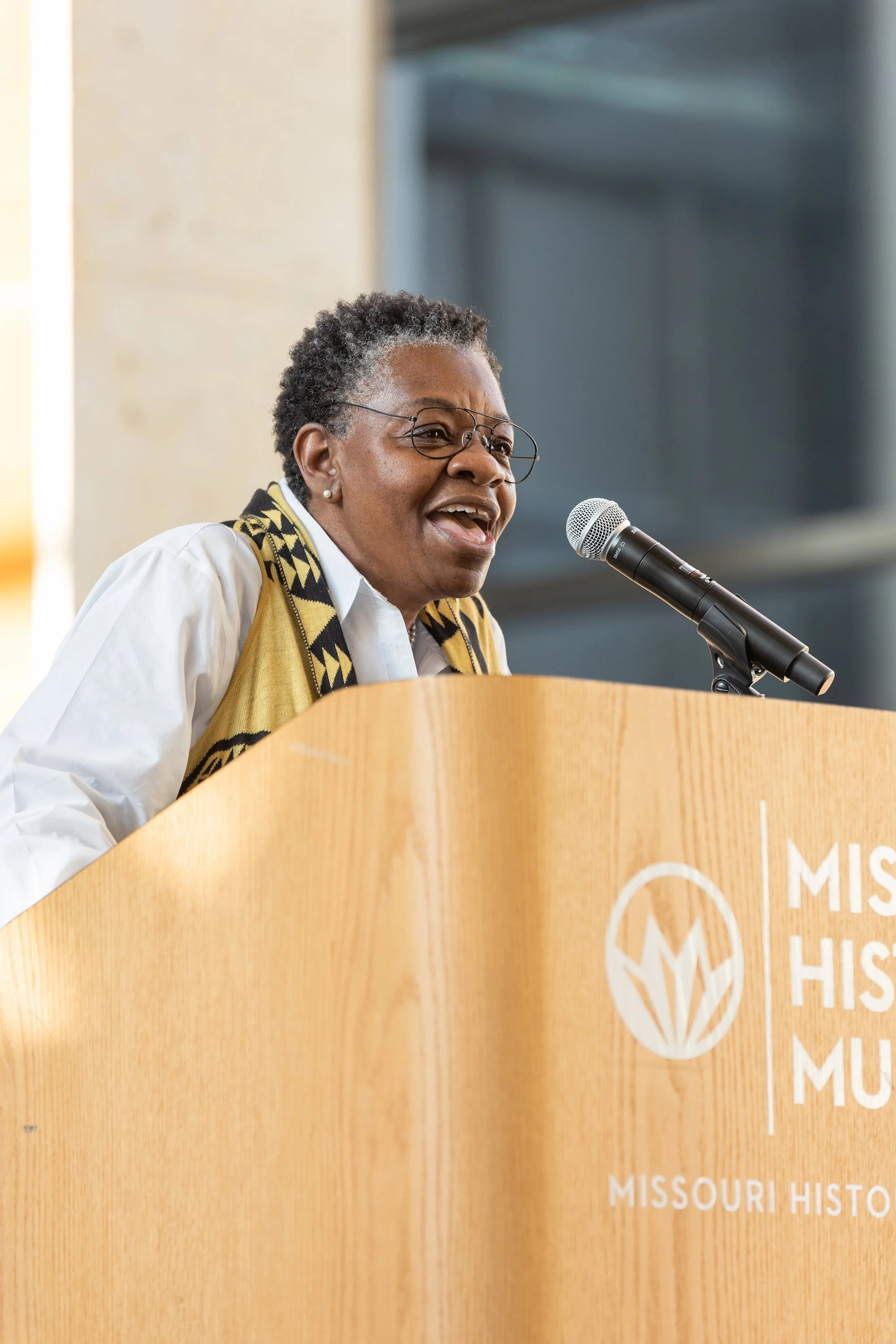 A woman speaking at a podium with a Missouri history event logo, wearing glasses, a white blouse, and a black and yellow scarf.
