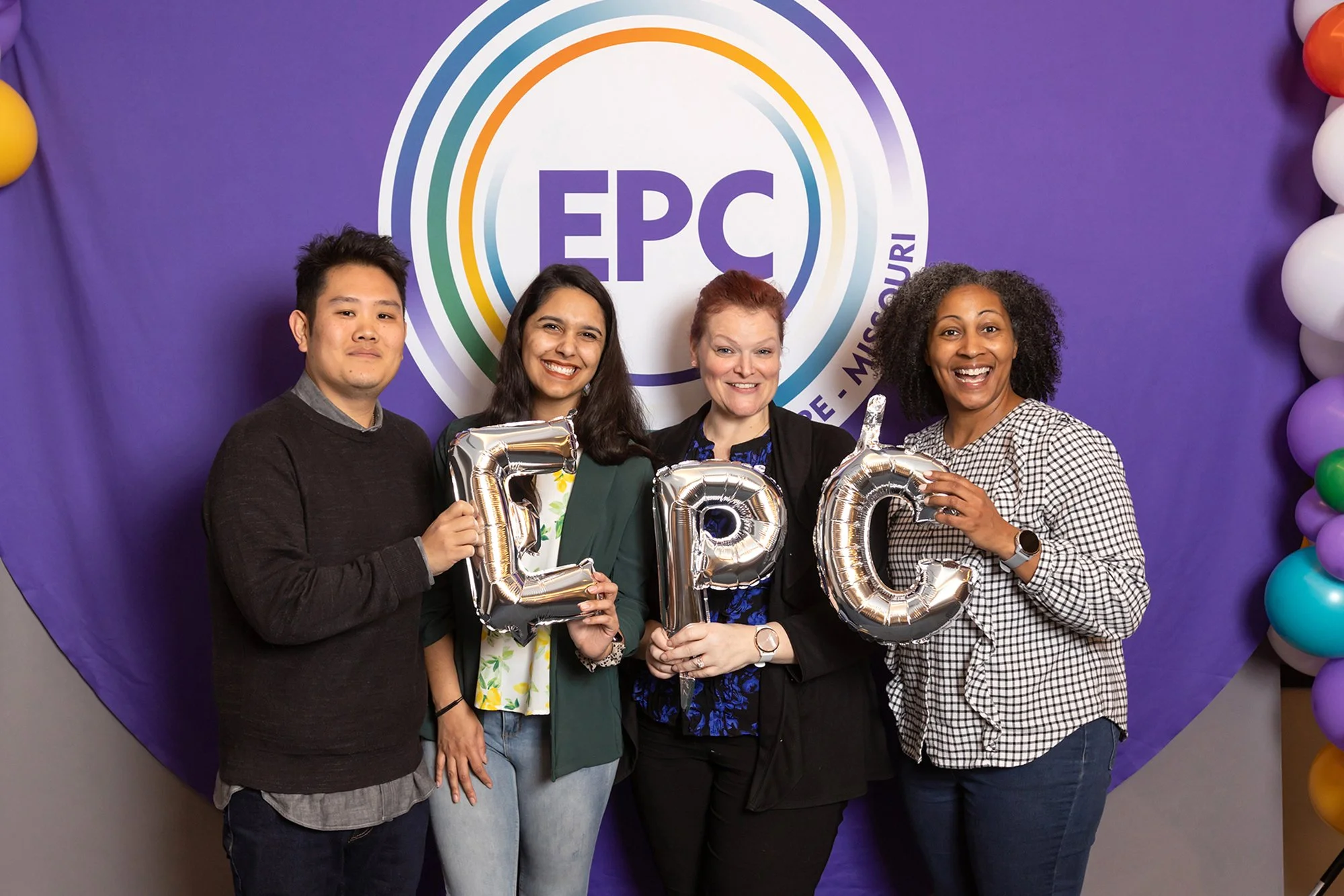 Four people standing together at an event, smiling and holding balloon letters spelling 'EPC,' in front of a purple backdrop with the 'EPC' logo and rainbow design.