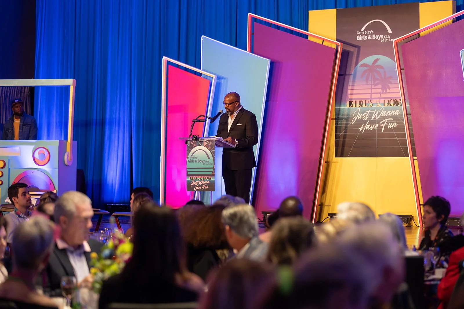 A man wearing a black suit and glasses is giving a speech at a podium decorated with a sign for the Girls & Boys Club of St. Louis. The background features colorful, oversized, tilted panels with neon lighting, and a blue curtain. A few people are seated at tables in the foreground, watching the speaker.