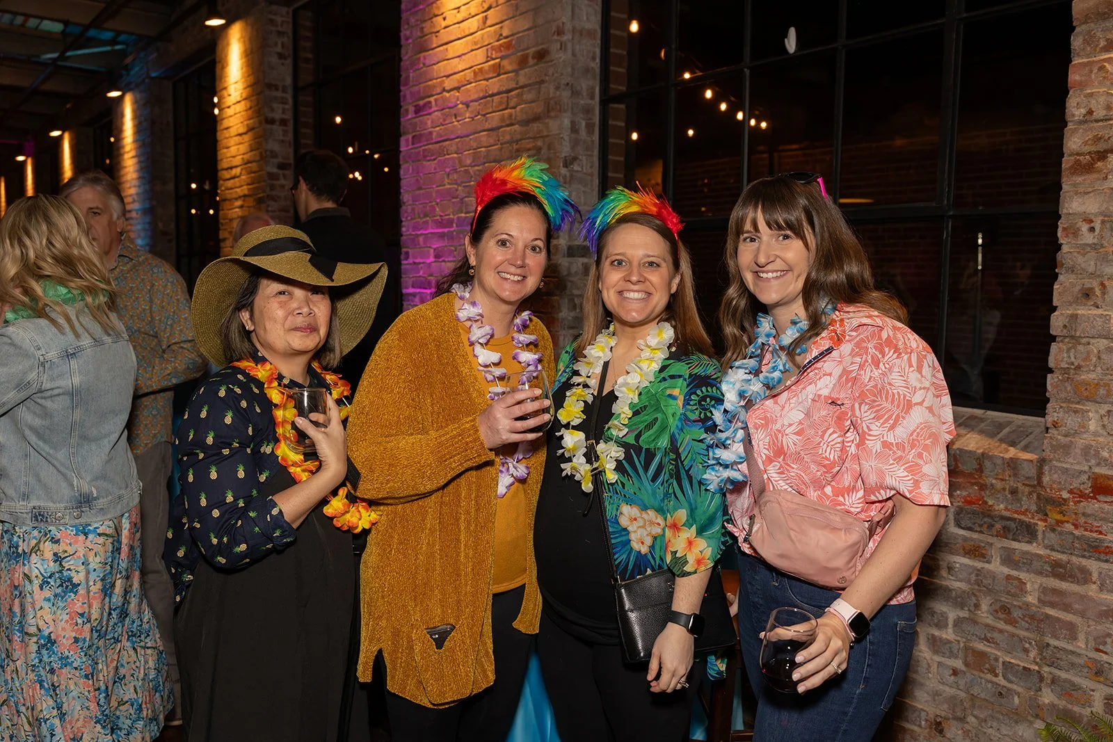 Four women at a party dressed in tropical and Hawaiian-themed attire, wearing leis and colorful accessories, smiling and holding drinks, in a festive indoor setting with brick walls and string lights.