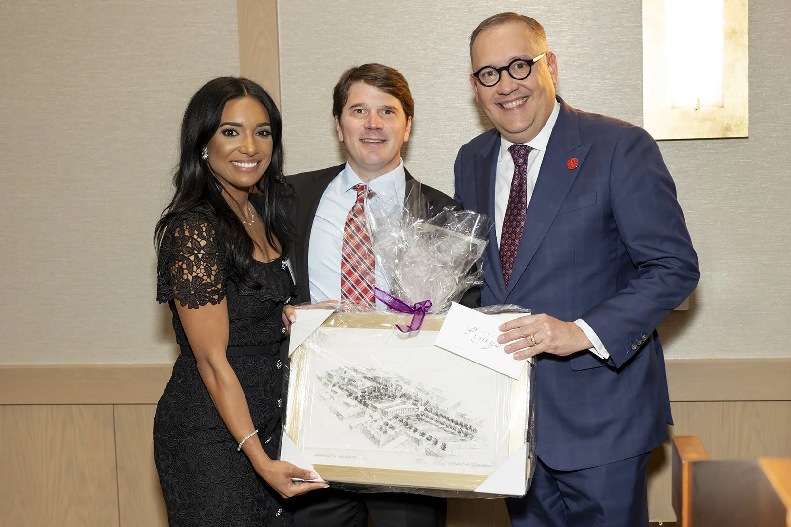 Three people standing together holding a framed drawing and a gift basket, smiling at the camera in a conference room.