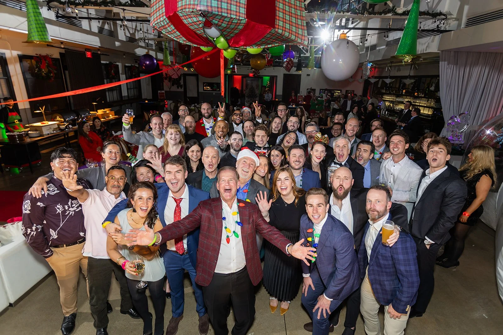 Group of people celebrating at a party with Christmas decorations, many smiling, some holding drinks, in a decorated venue.