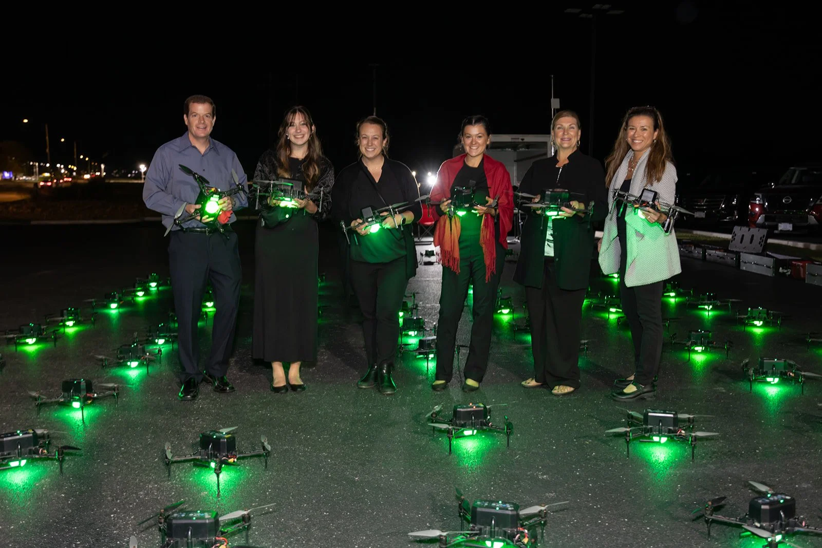 Group of seven people standing in a parking lot at night holding drones with green lights, with more drones on the ground around them.