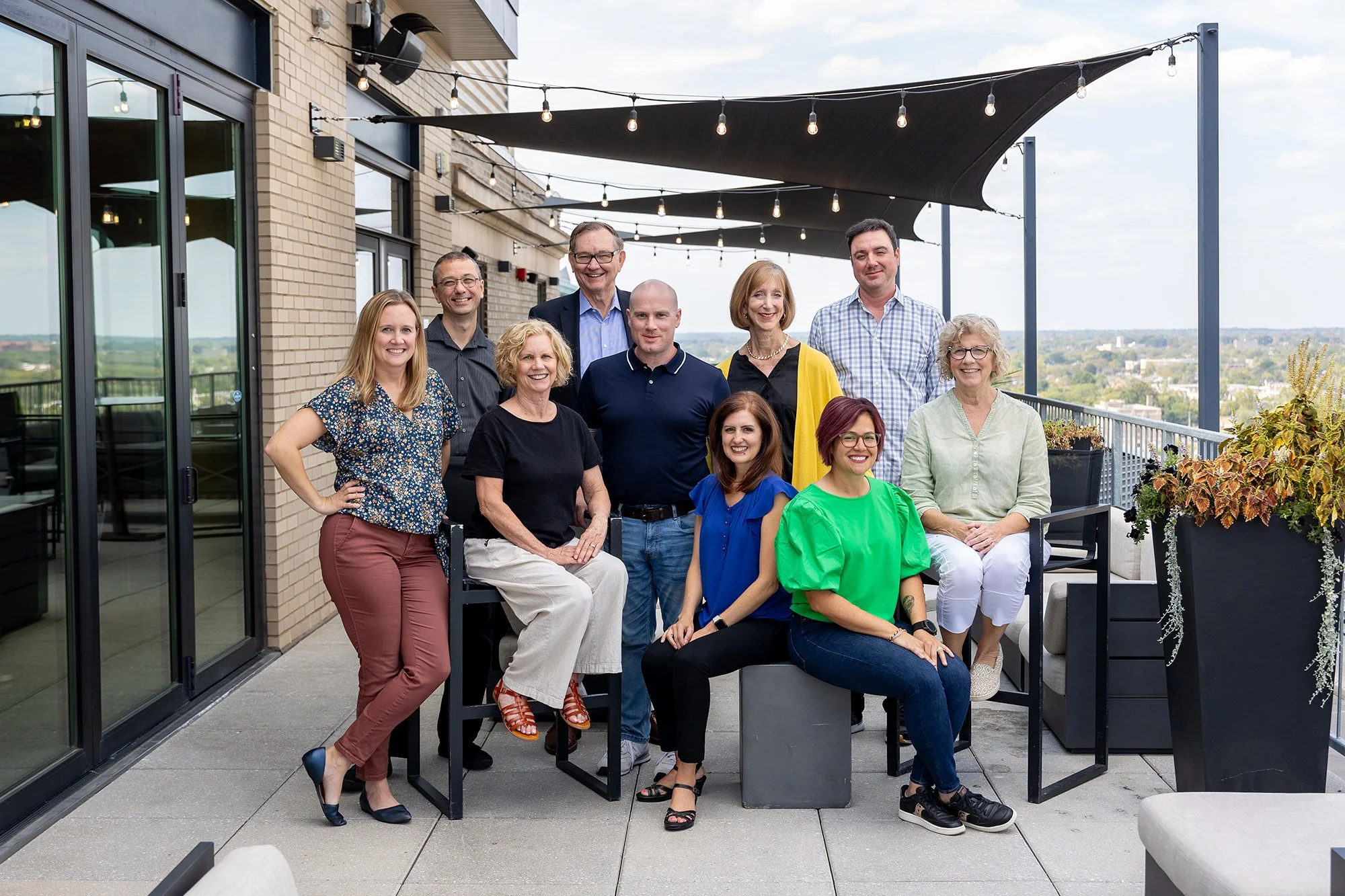 A group of eleven diverse adults posing on a rooftop terrace with glass doors, string lights, and a view of the city and landscape in the background.