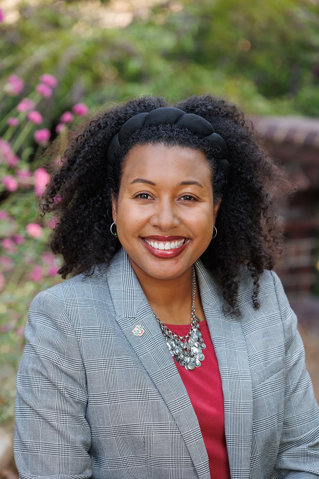 A woman with curly black hair, wearing a checkered blazer, red top, and a silver necklace, smiling outdoors with pink flowers and greenery in the background.