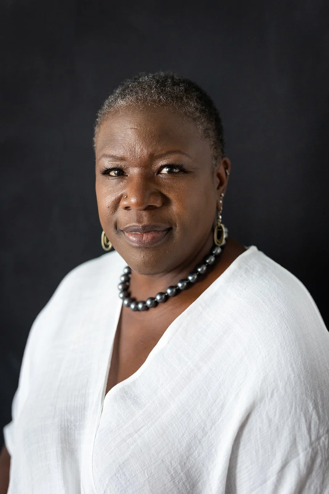 Portrait of a woman with short, curly gray hair, wearing a white blouse, pearl necklace, and earrings, against a black background.