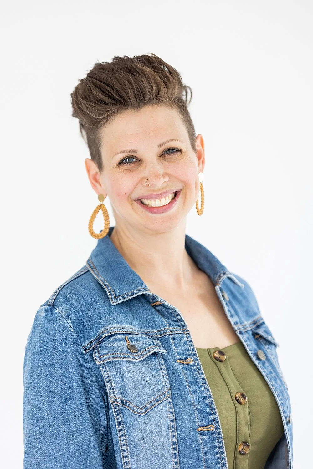 A woman with short brown hair styled up, wearing large oval earrings, a denim jacket, and a green top, smiling against a white background.