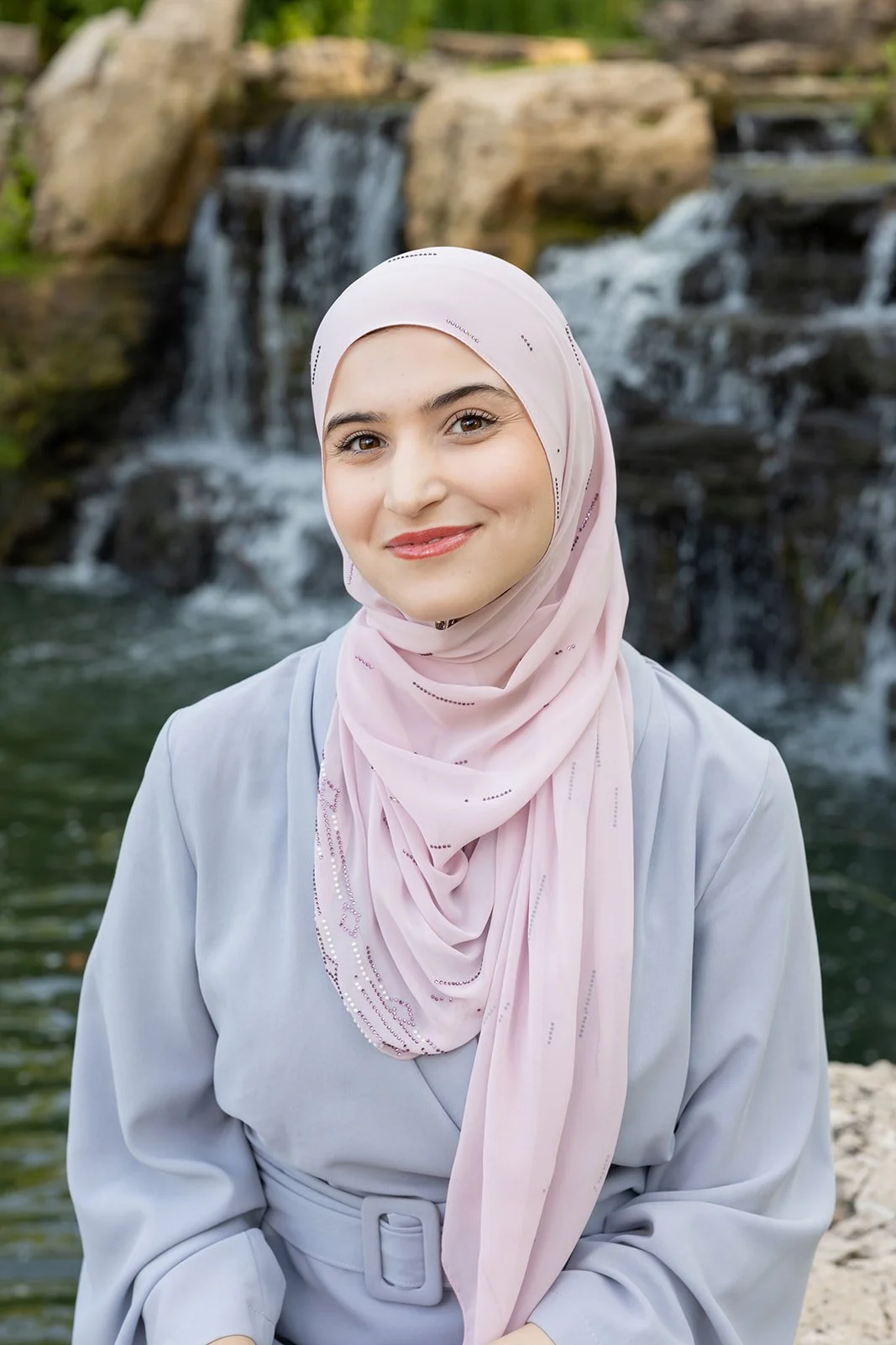 A woman wearing a hijab standing near a waterfall with rocks and greenery in the background.
