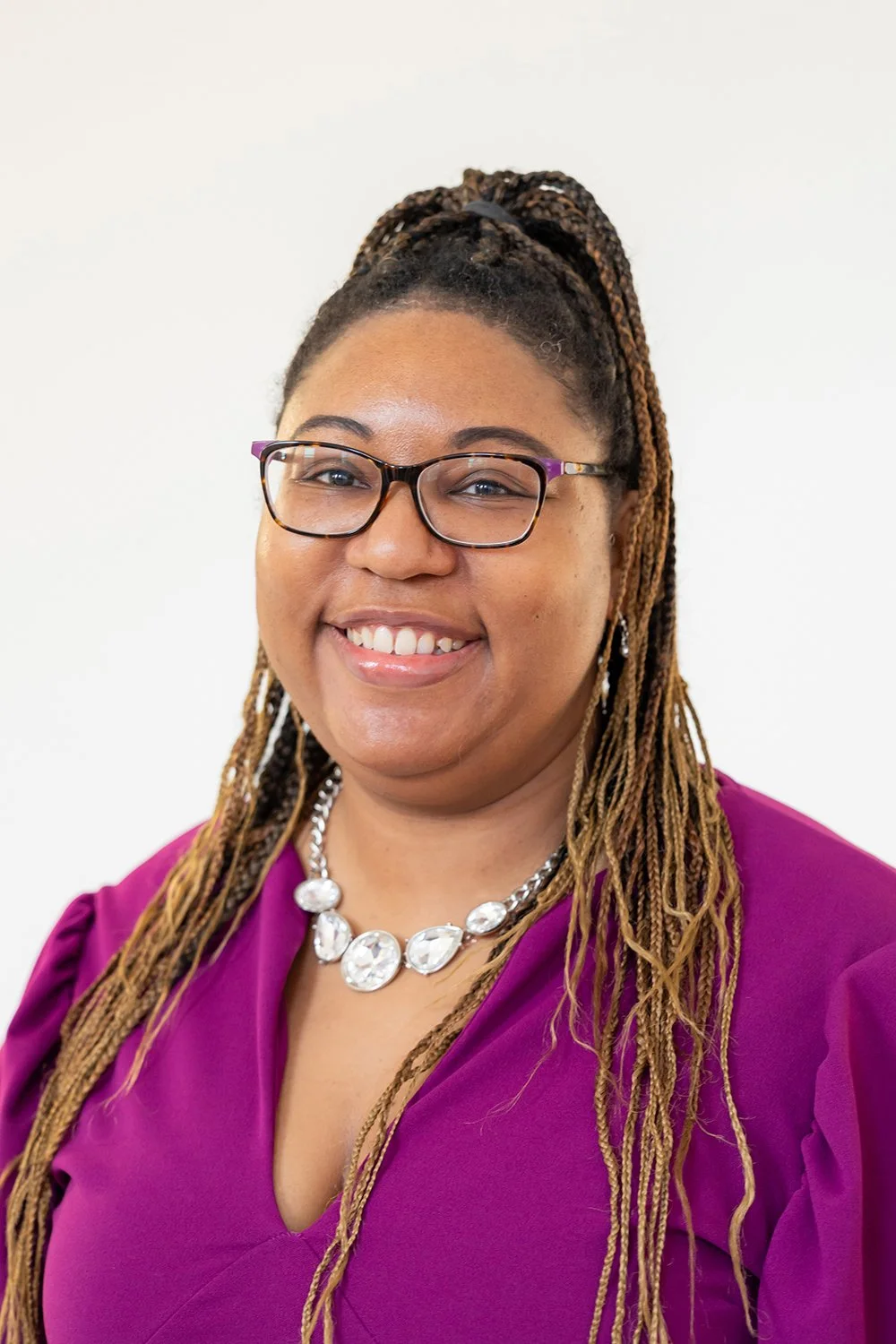 A woman with glasses and braided hair, smiling, wearing a purple blouse and a statement necklace, against a white background.