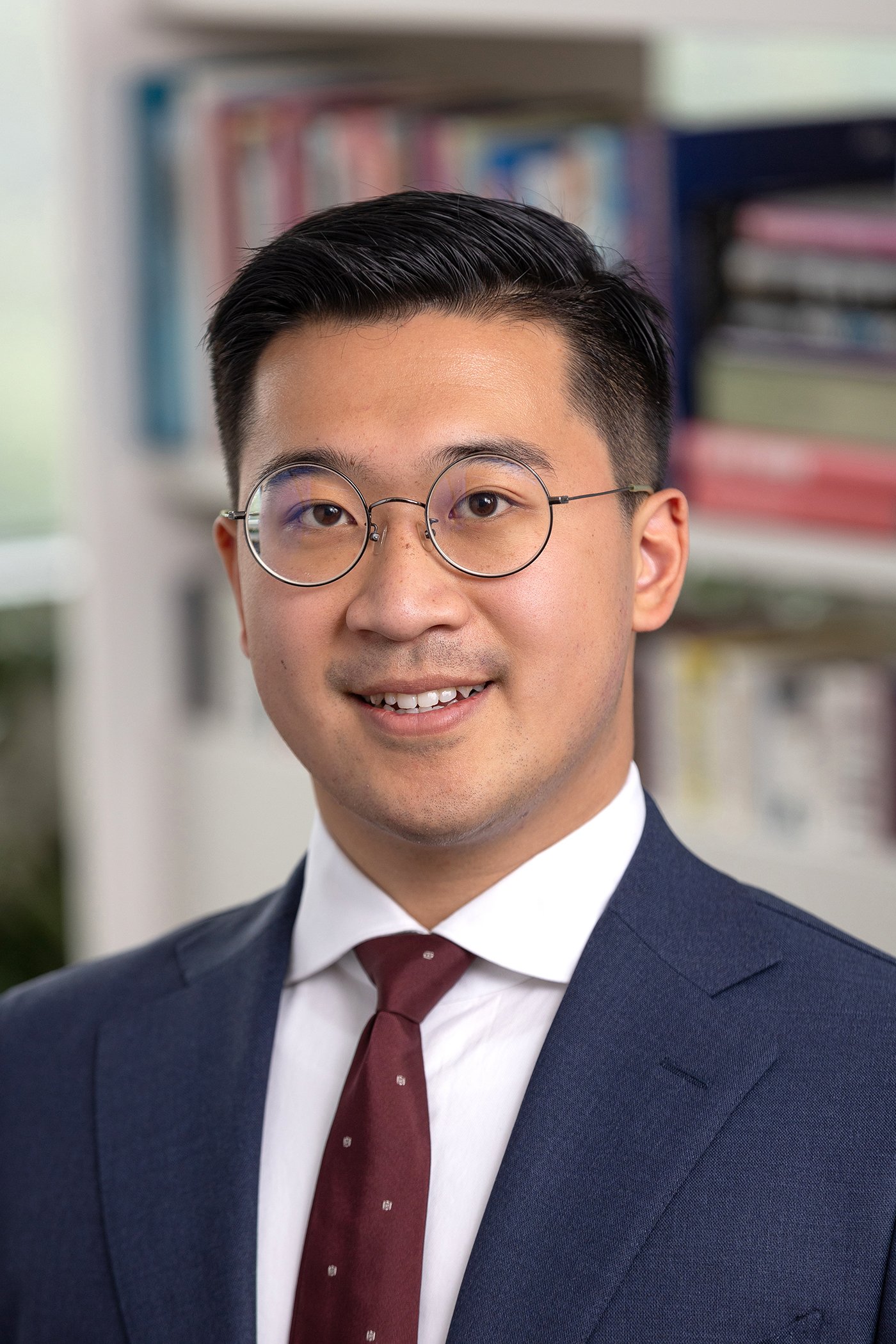 A professional young man wearing glasses, a navy suit, a white shirt, and a maroon tie, smiling in an office setting with bookshelves in the background.