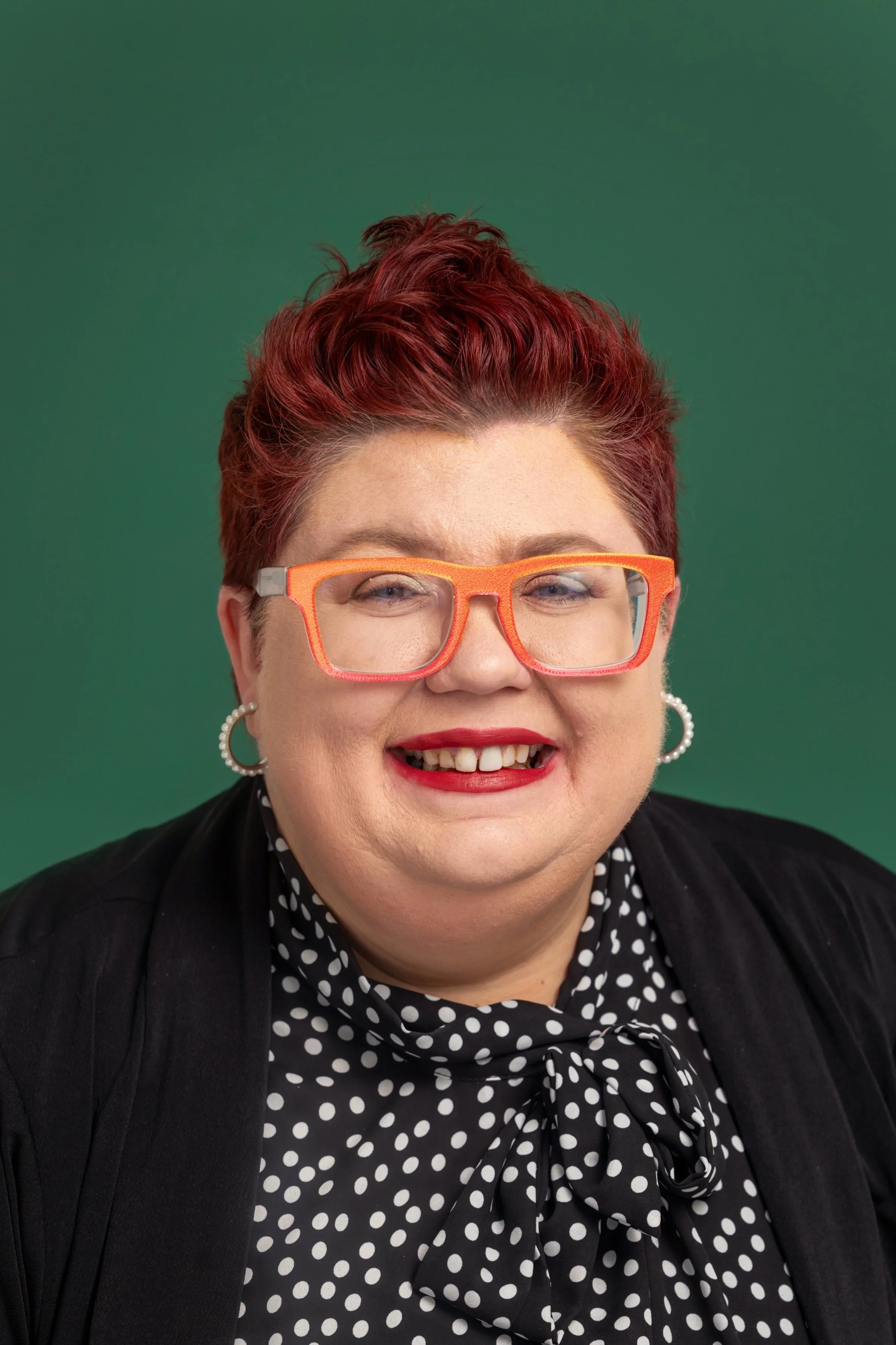 Woman with short red hair, orange glasses, pearl earrings, polka dot blouse with a bow, black jacket, smiling against a green background.
