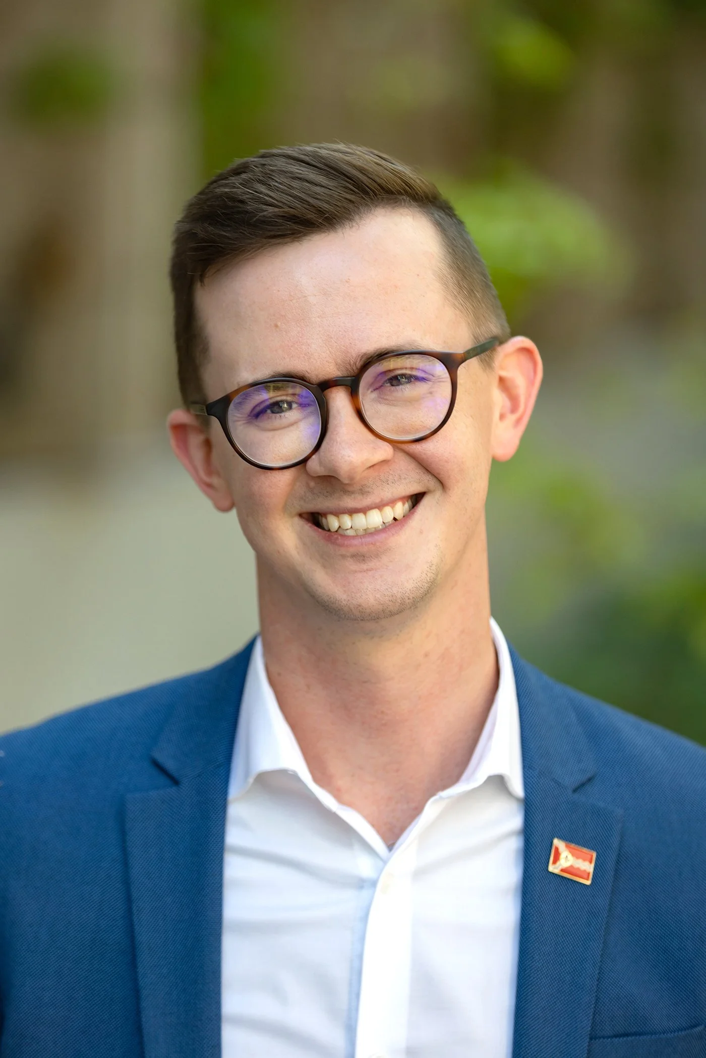 A smiling young man wearing glasses, a white shirt, and a blue blazer with a pin, standing outdoors with a blurred background of trees and greenery.