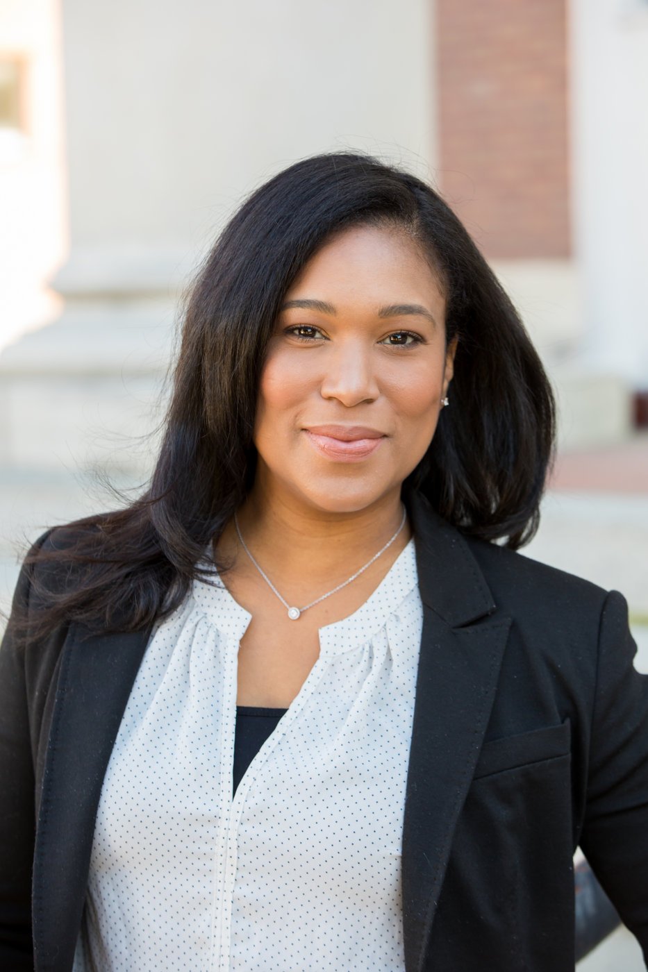 A professional woman with black hair wearing a black blazer and white blouse standing outdoors.