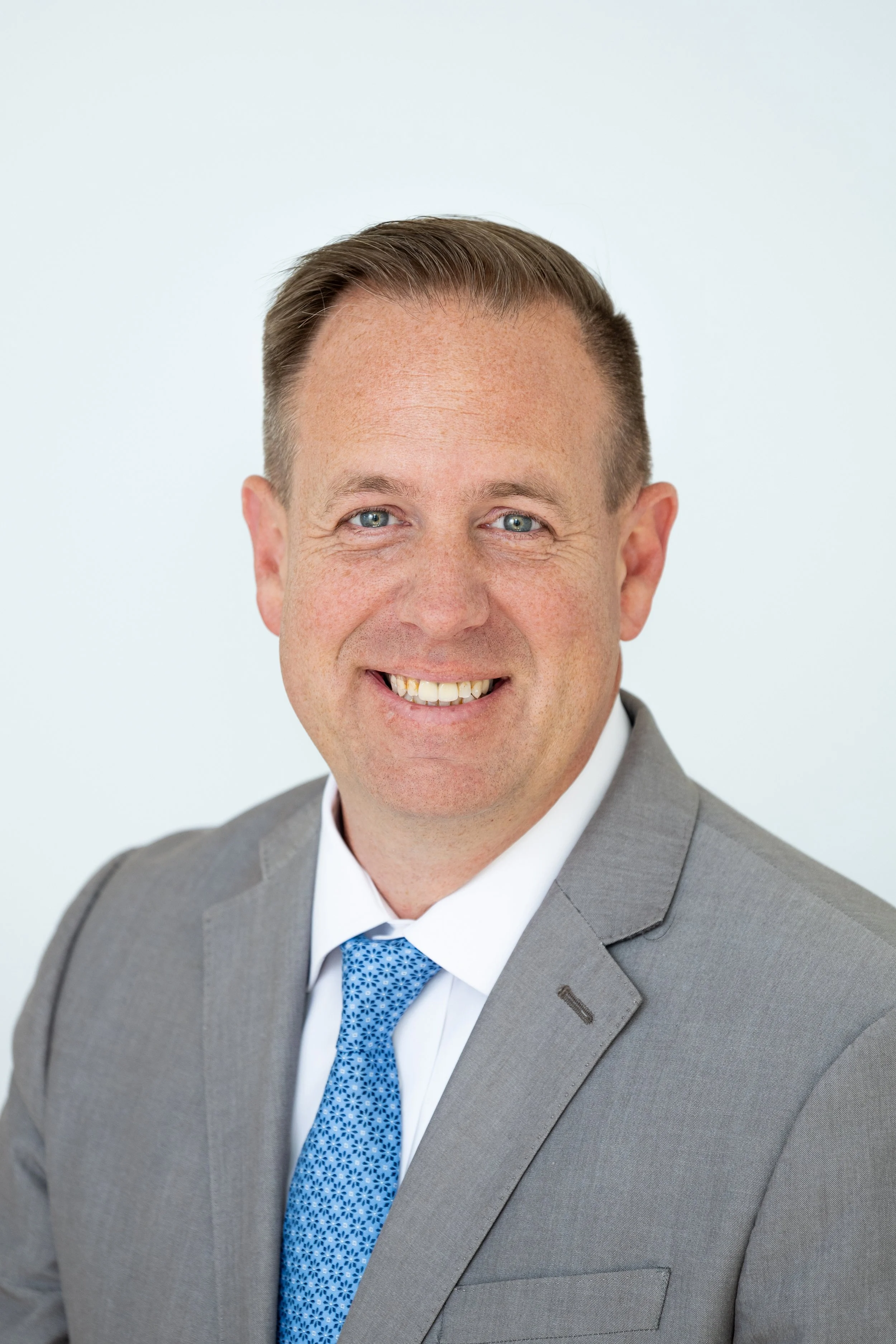 A man in a gray suit with a white shirt and blue patterned tie, smiling at the camera, with a plain light background.