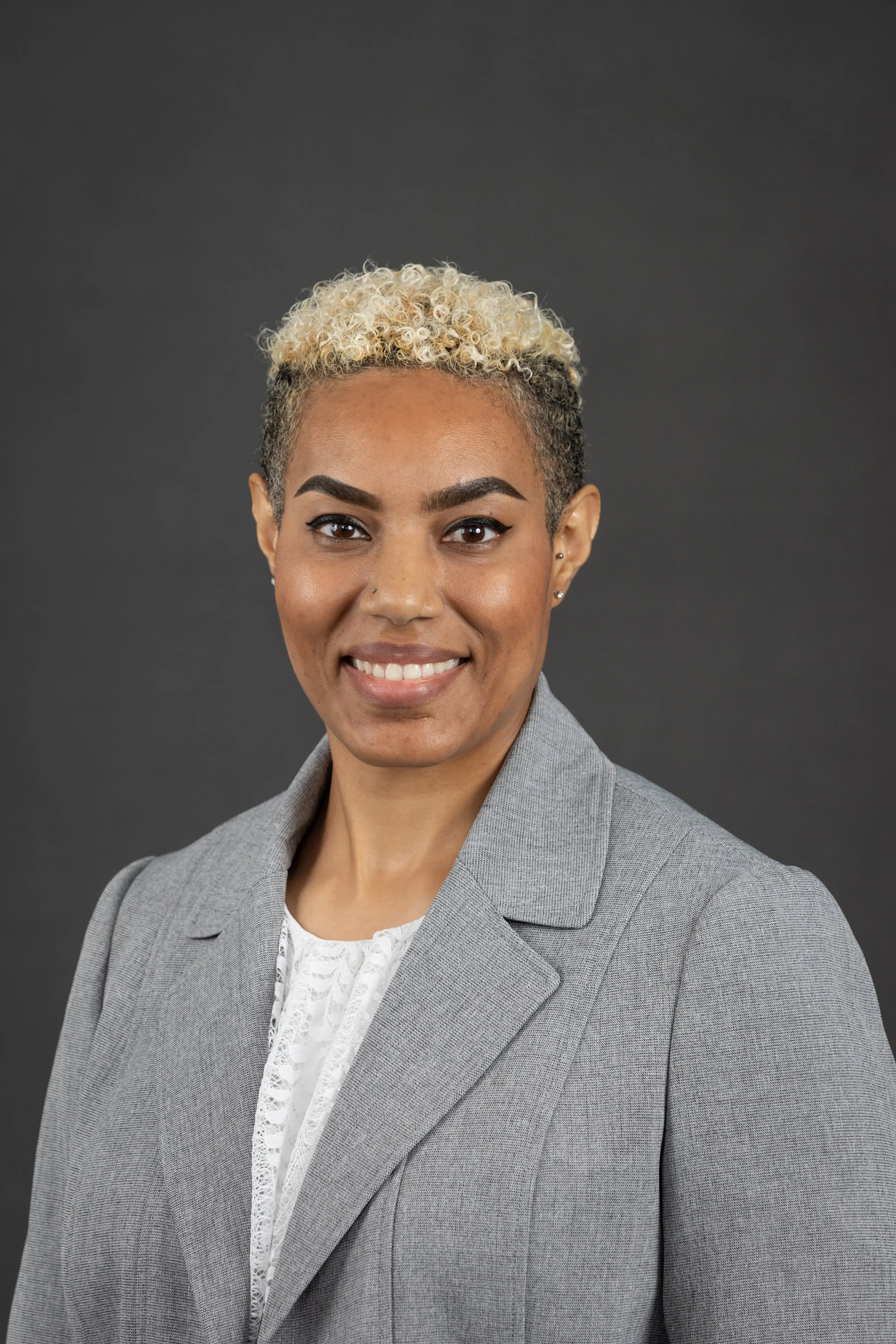 A professional woman with short, curlyblonde hair, wearing a gray blazer and a white lace blouse, smiling against a dark gray background.