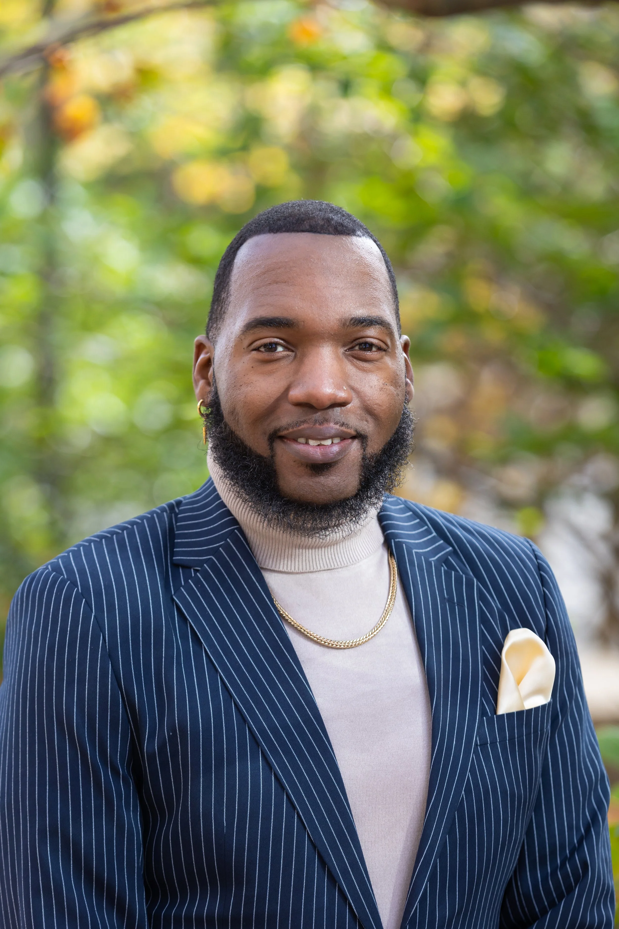 A man with a beard and earrings, wearing a striped blazer, a beige turtleneck, a gold chain necklace, and a pocket square, standing outdoors with green foliage in the background.