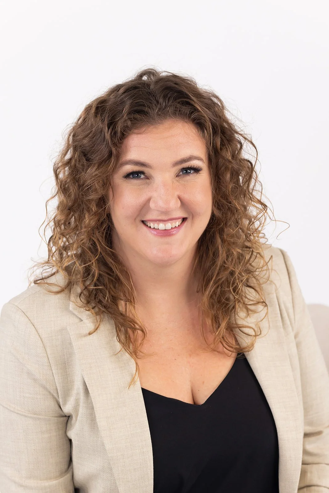 Smiling woman with curly hair wearing a beige blazer and black top against a white background.